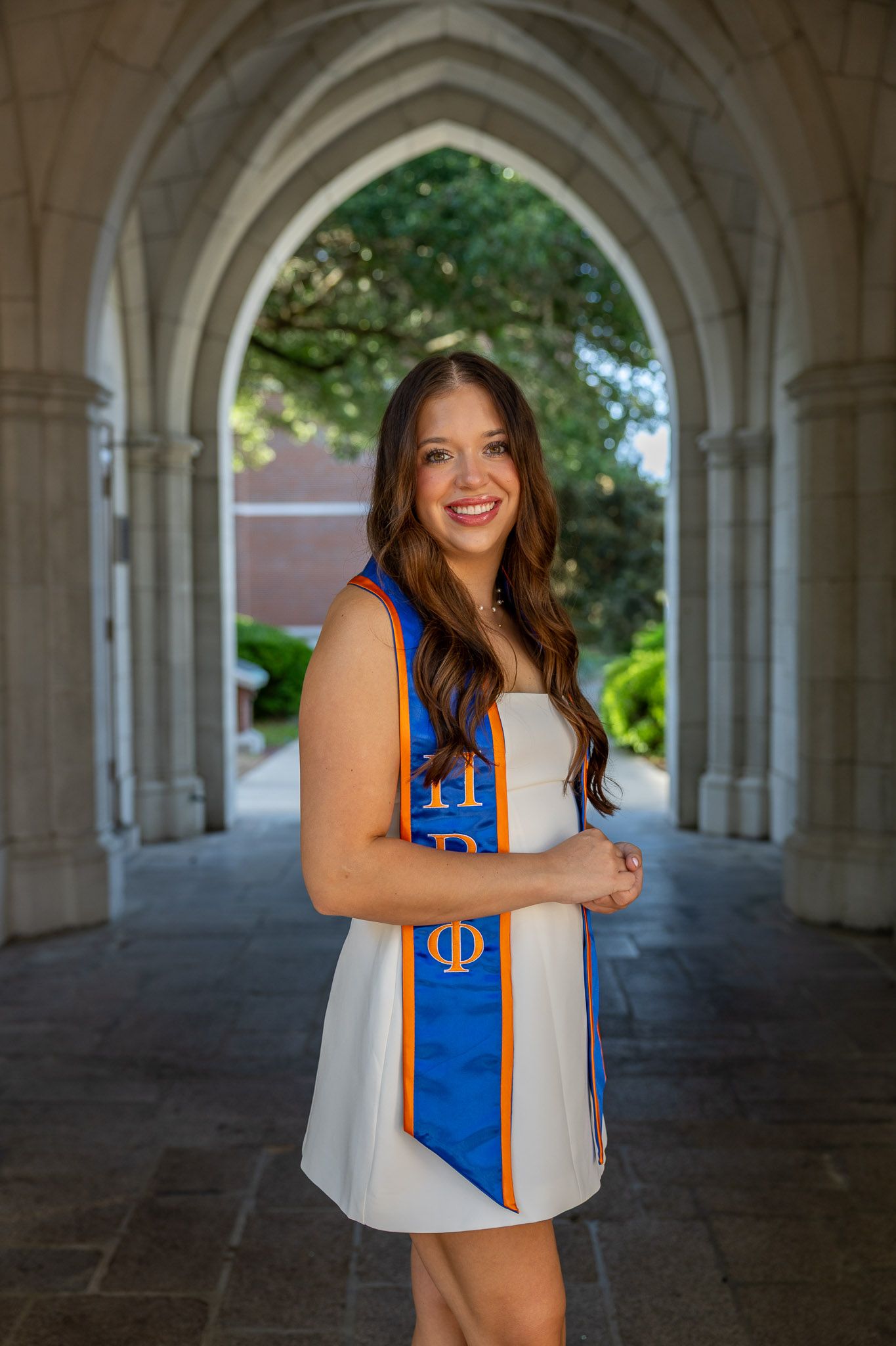 Smiling young woman in white dress with blue and orange graduation stole posing under stone arches on college campus.