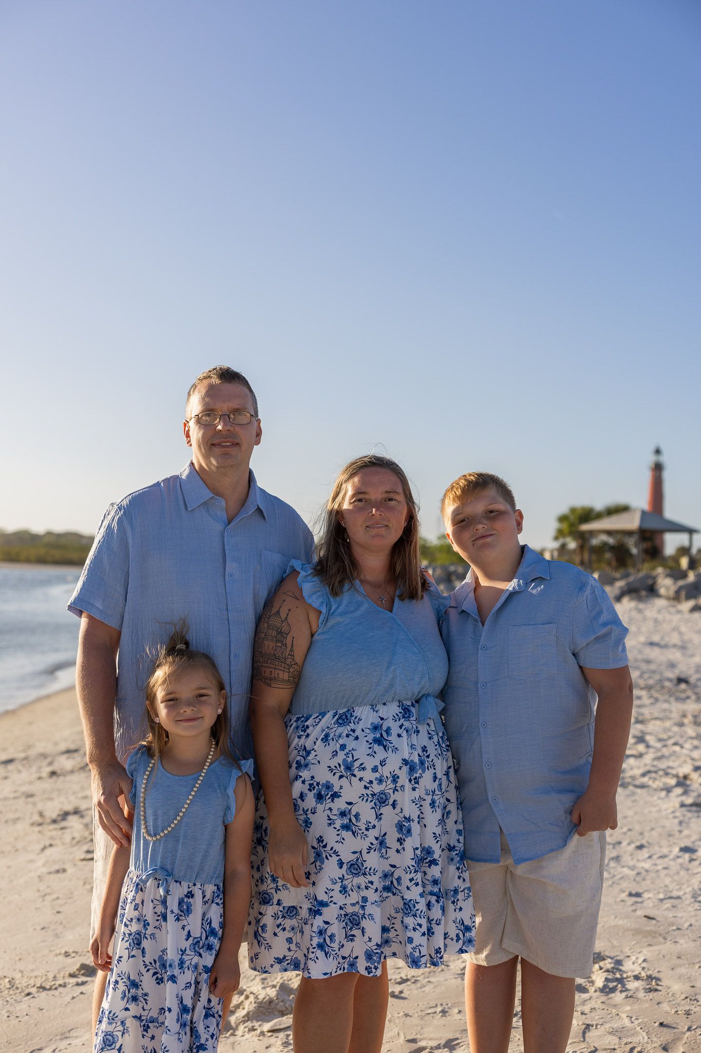 Family of four posing on a sunny beach near a lighthouse, wearing coordinating blue and white outfits.