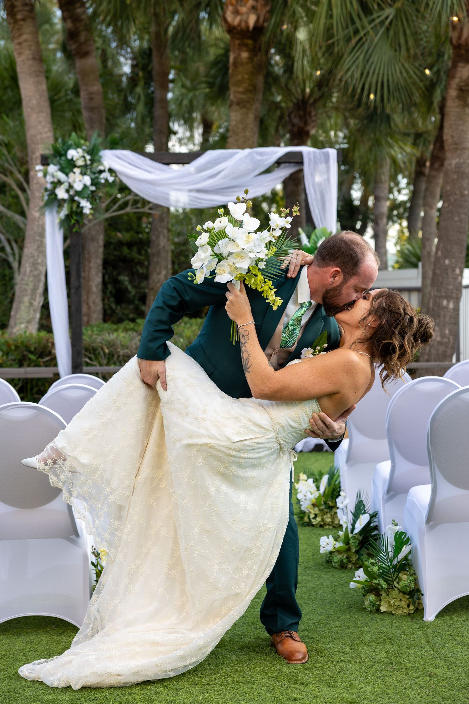 A romantic dip pose during a wedding ceremony with white chairs and palm trees.