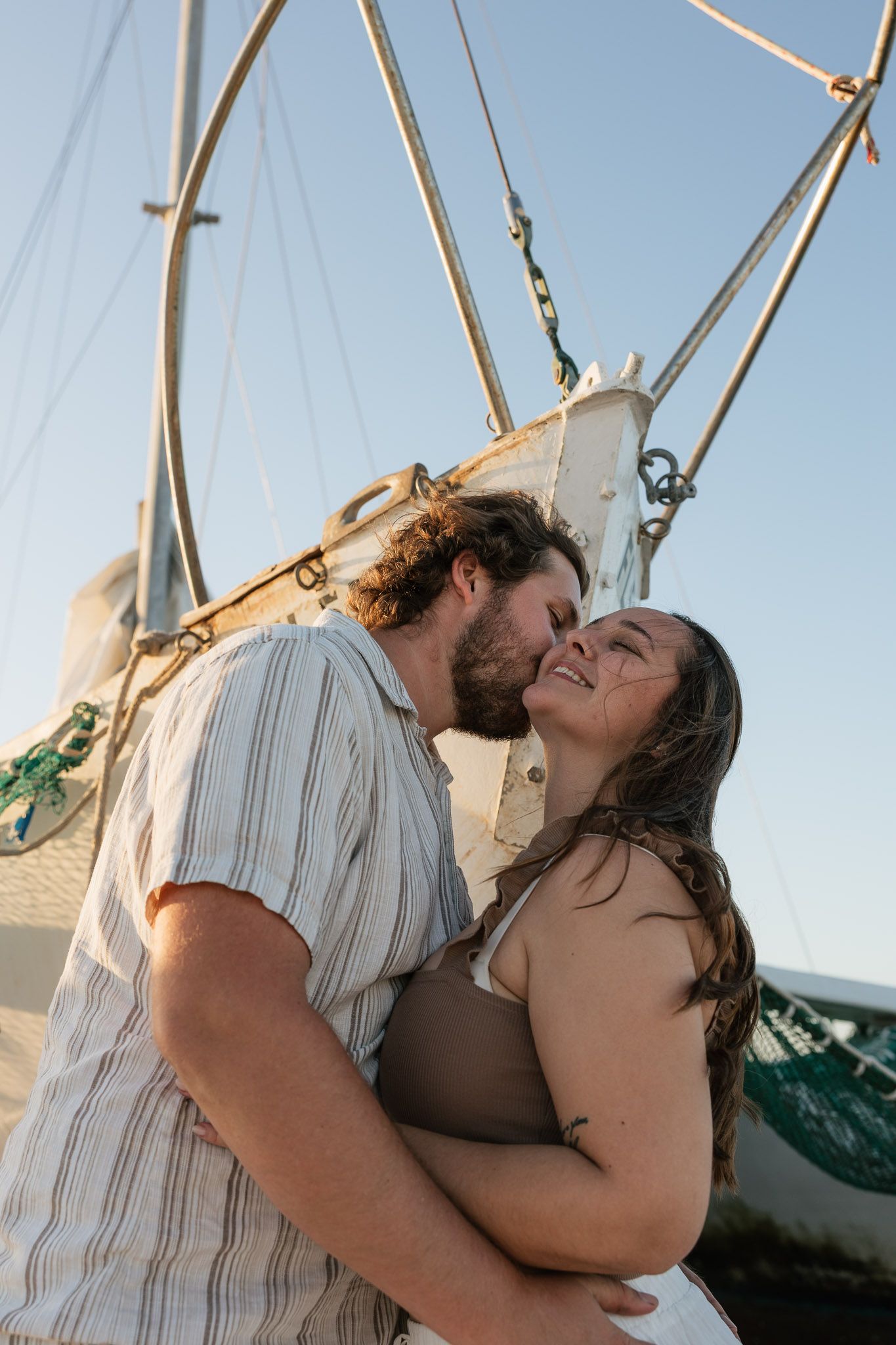 A couple shares a romantic kiss near a sailboat mast against a clear blue sky at golden hour.