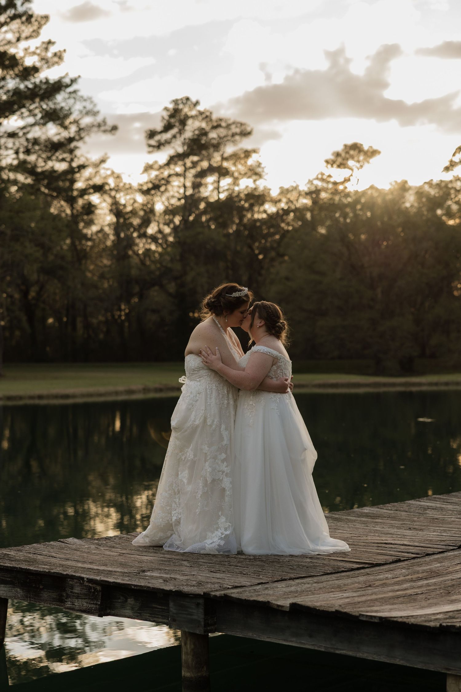 Two brides in white gowns share a romantic moment on a wooden dock by a lake at sunset.