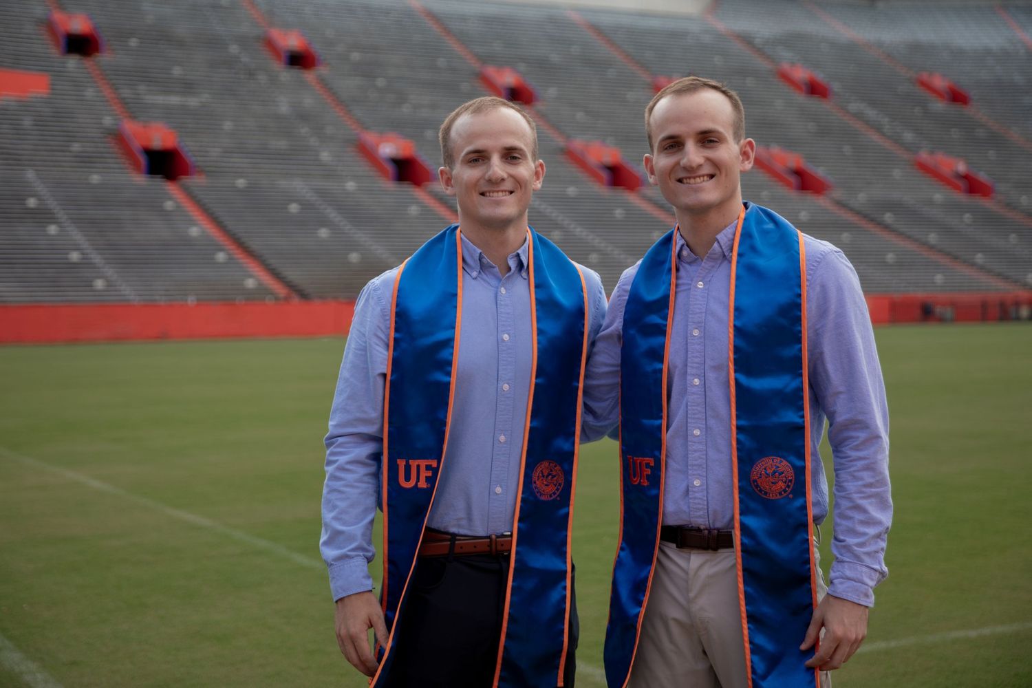 Two graduates wearing blue stoles stand on football field with stadium seats in background.