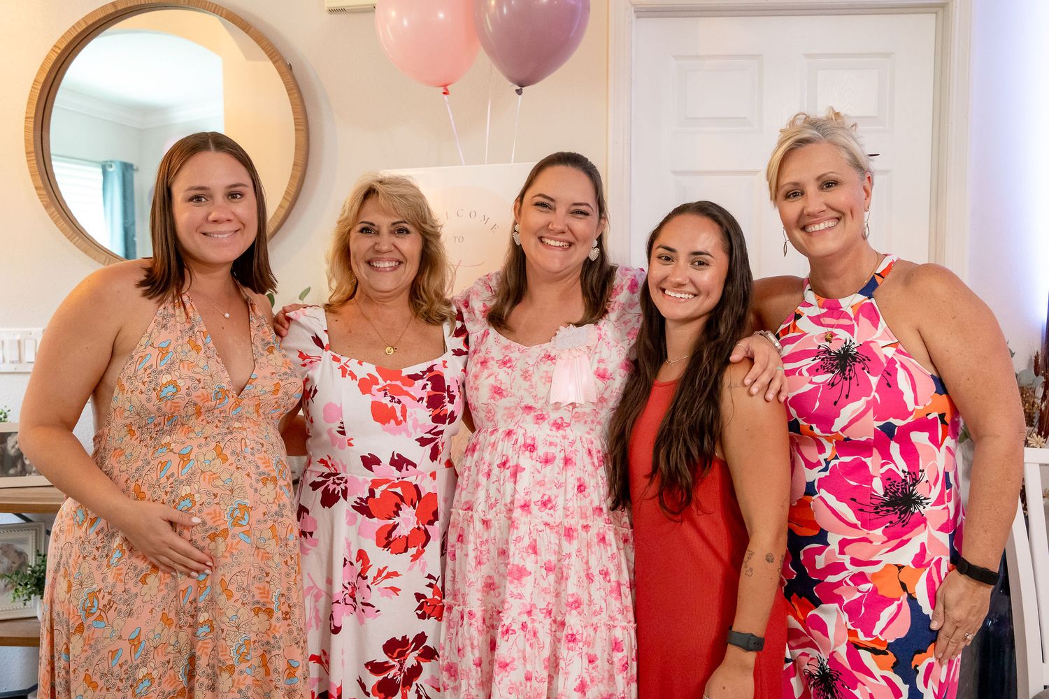 Group of women pose together at a party wearing floral pink and coral dresses.
