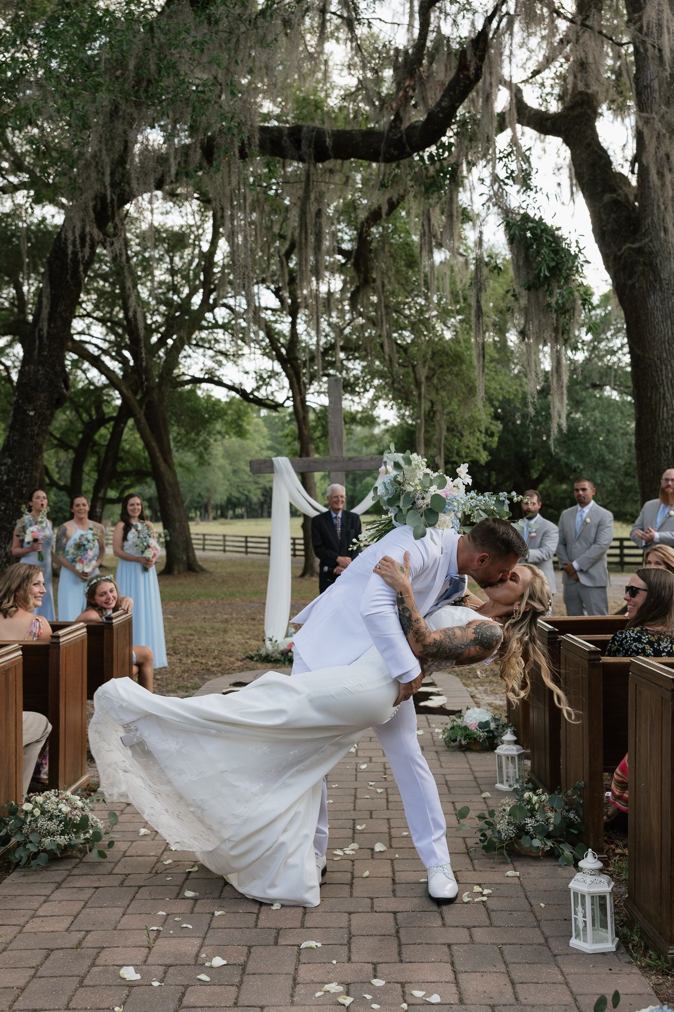 Groom dips bride for a kiss at outdoor wedding ceremony with Spanish moss trees and bridesmaids in blue.