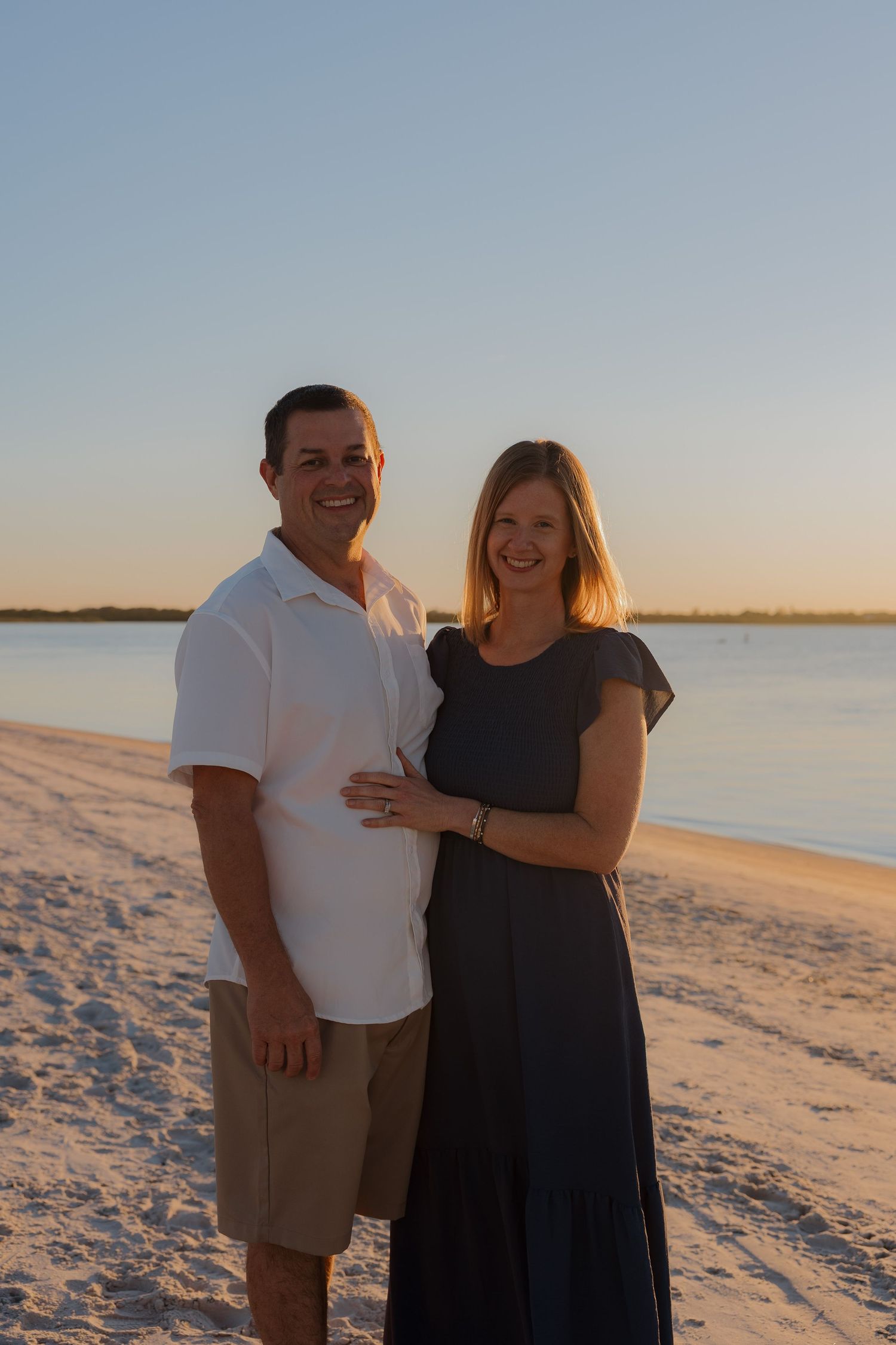 Couple in casual attire embracing on white sandy beach at sunset.