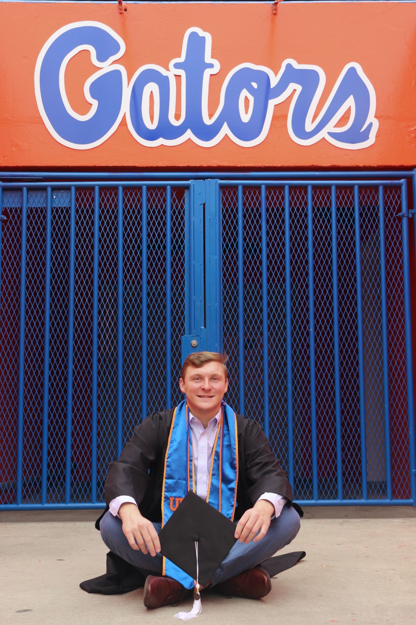 Graduate poses beneath orange Gators sign against blue metal grating background.