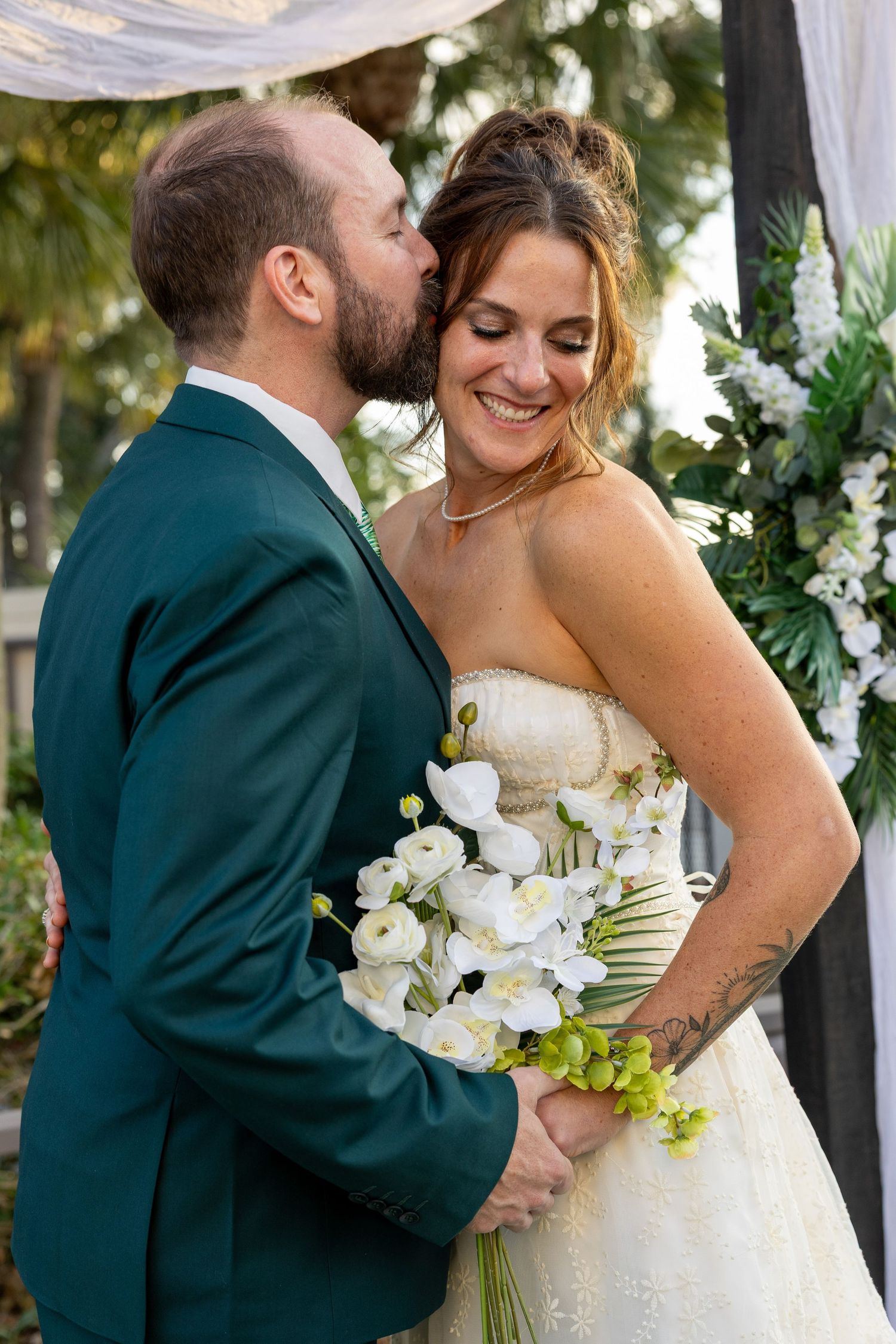 Romantic wedding couple embraces outdoors with white floral bouquet and green suit details.