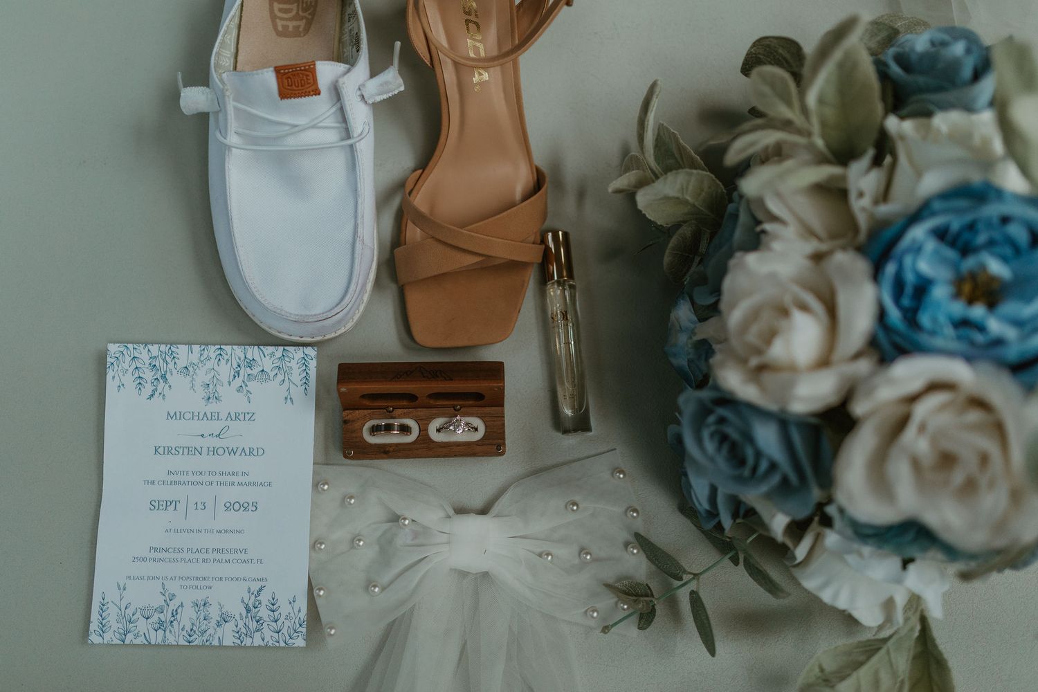 Wedding details including rings, invitations and blue floral arrangements arranged on a table.
