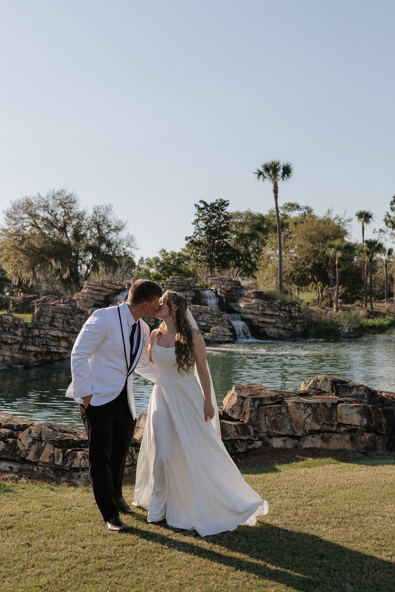 A bride and groom share a kiss outdoors near a scenic pond with a waterfall and palm trees on their wedding day.