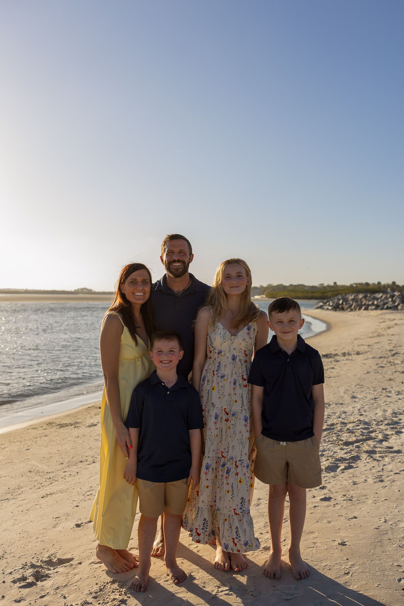 Family of five posing together on a sandy beach at sunset, wearing coordinated outfits in warm golden light.