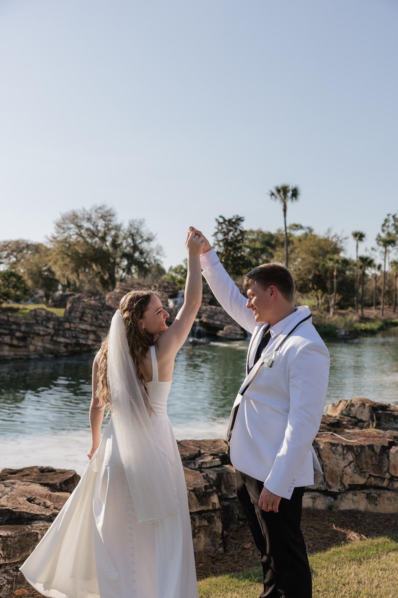 Bride and groom dancing outdoors near a scenic waterway, bride in white gown with veil, groom in white tuxedo jacket.