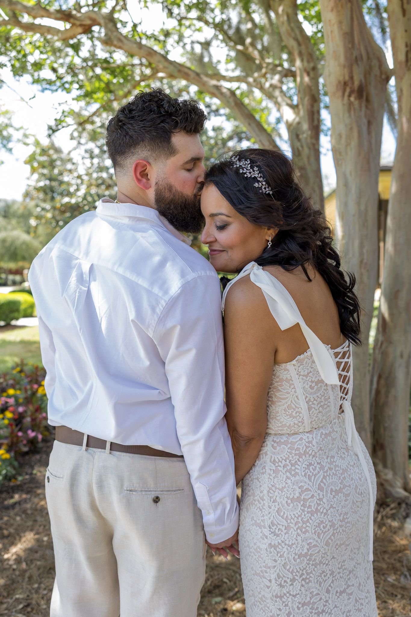 A groom in white shirt kisses his bride wearing a lace wedding dress and floral hair piece outdoors.