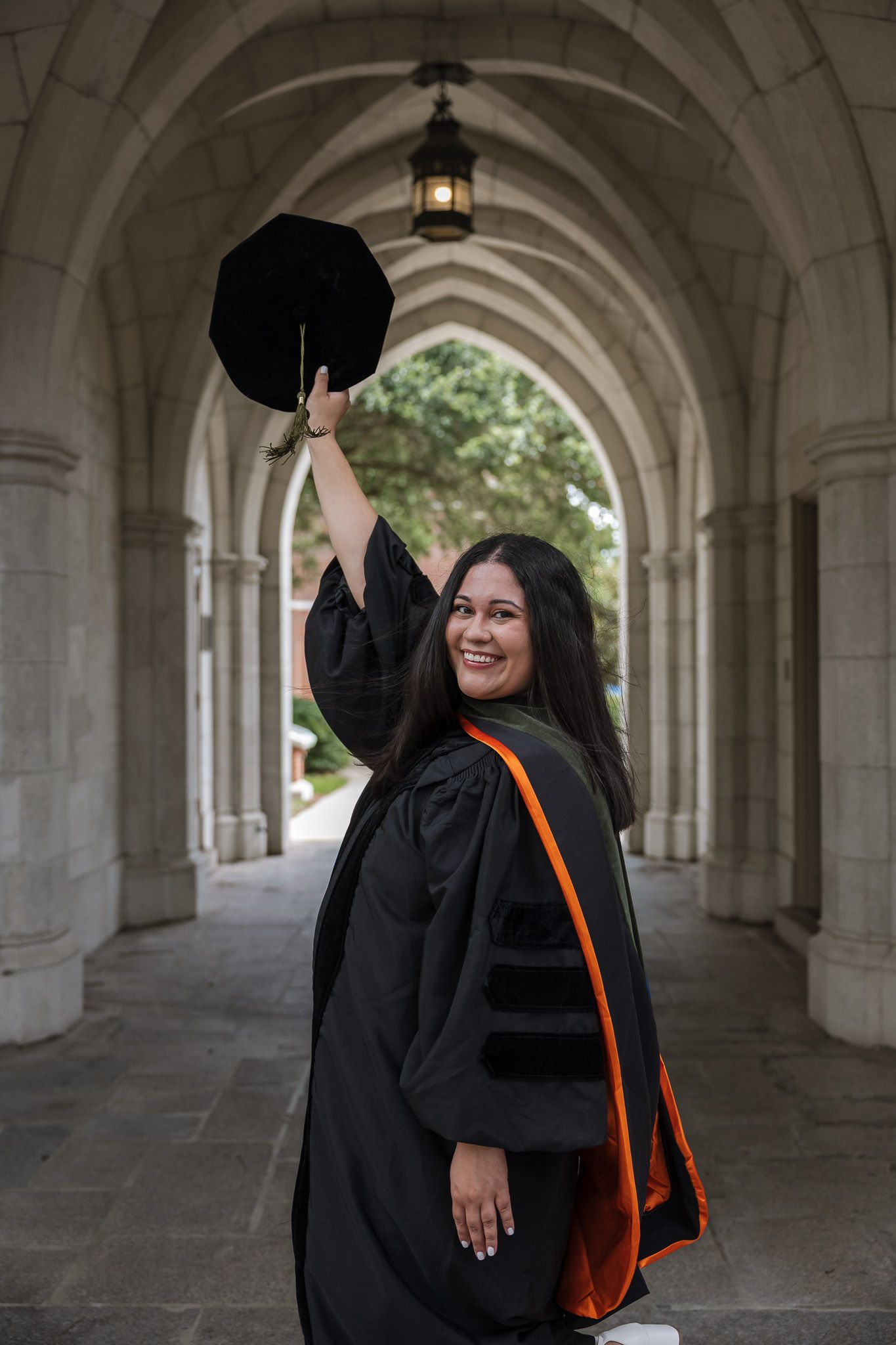 A smiling woman in black doctoral graduation gown with orange sash raises her cap under stone archway.