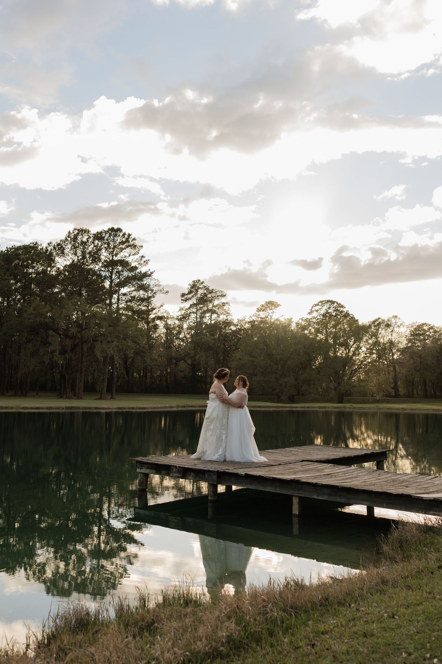 Two brides in white gowns embrace on a wooden dock overlooking a serene lake at golden hour.