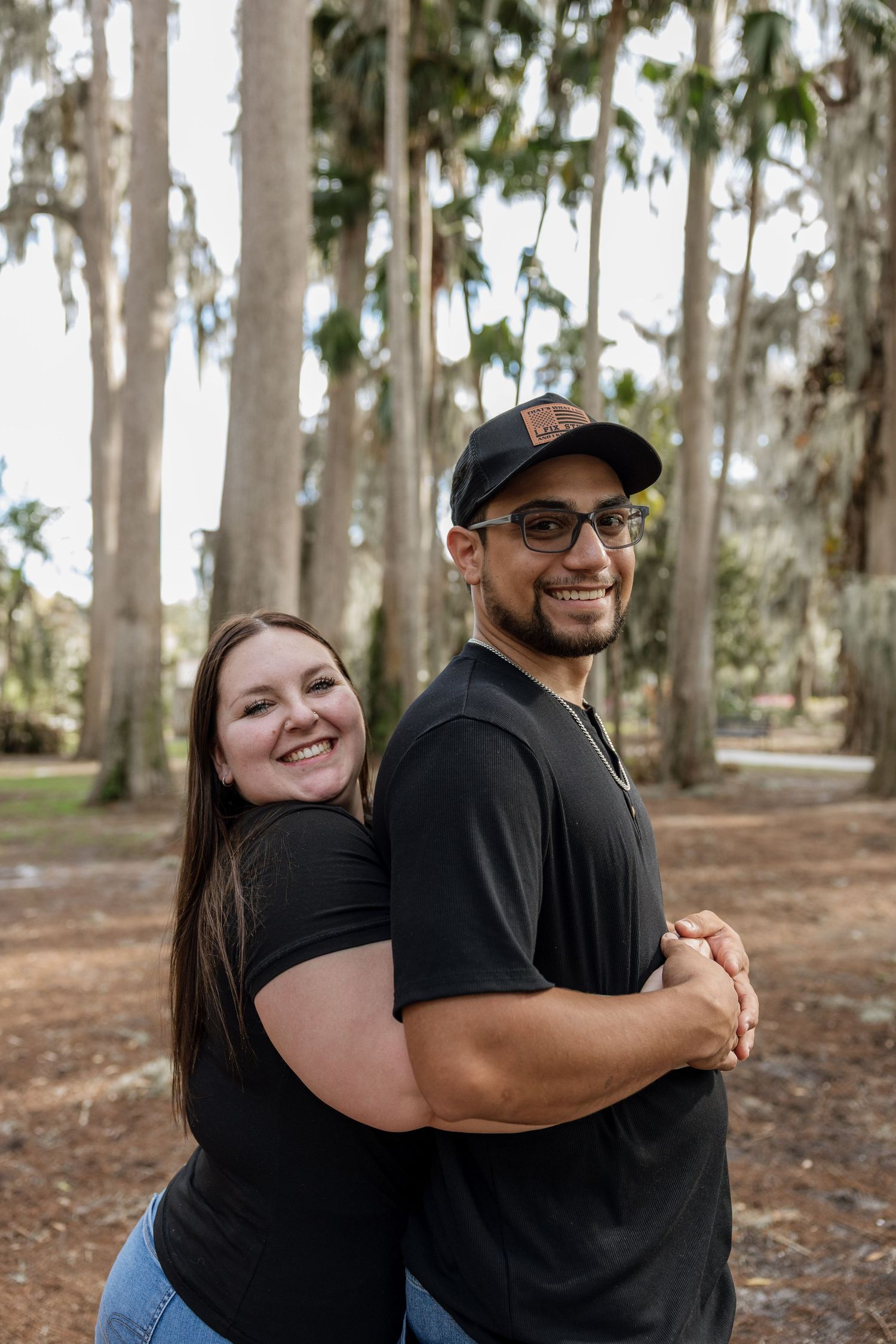 A couple embraces and smiles while standing among tall eucalyptus trees in a wooded area.