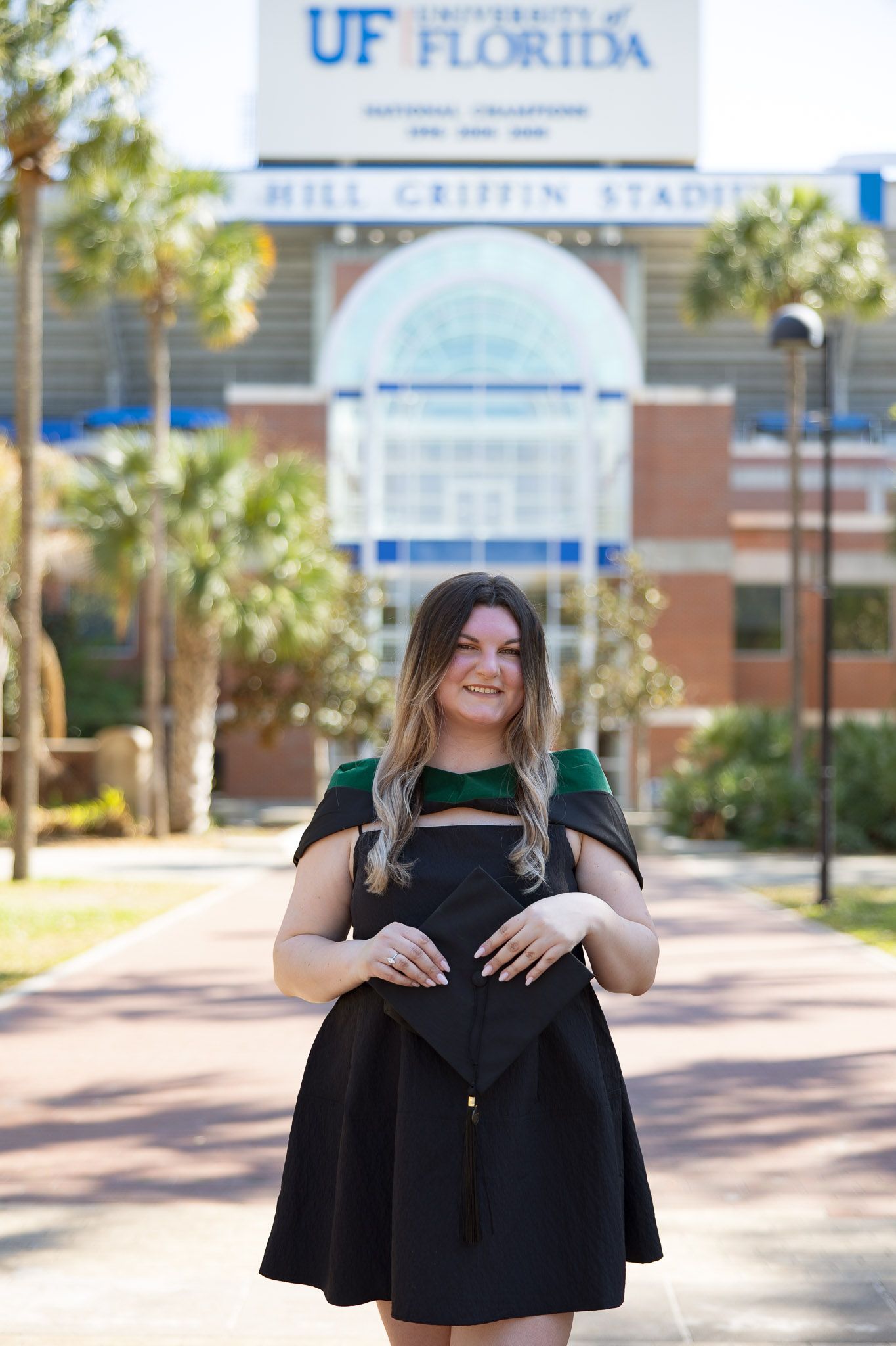 University of Florida graduate smiling in black dress holding cap in front of Ben Hill Griffin Stadium.