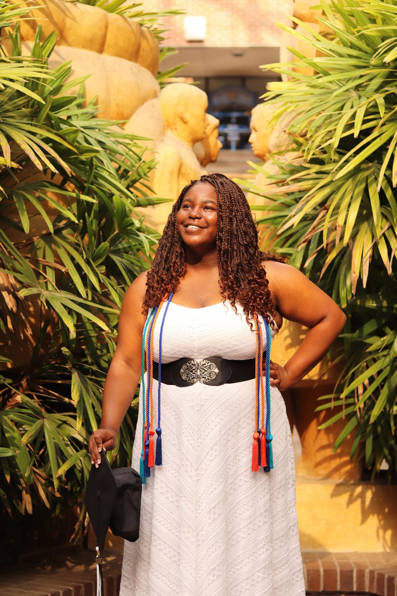 Graduate in white dress and academic regalia poses confidently among tropical foliage.