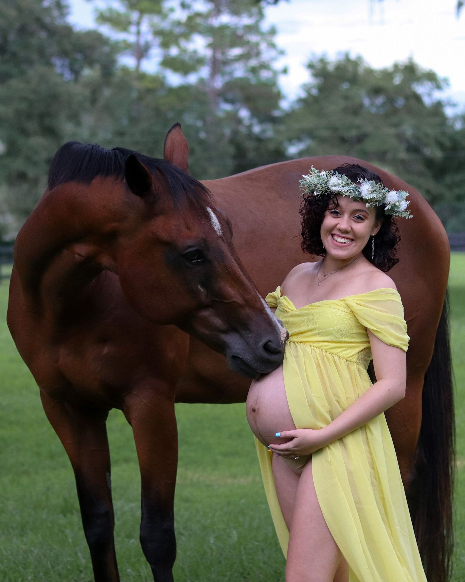 Pregnant woman in yellow dress shares moment with curious brown horse in field.