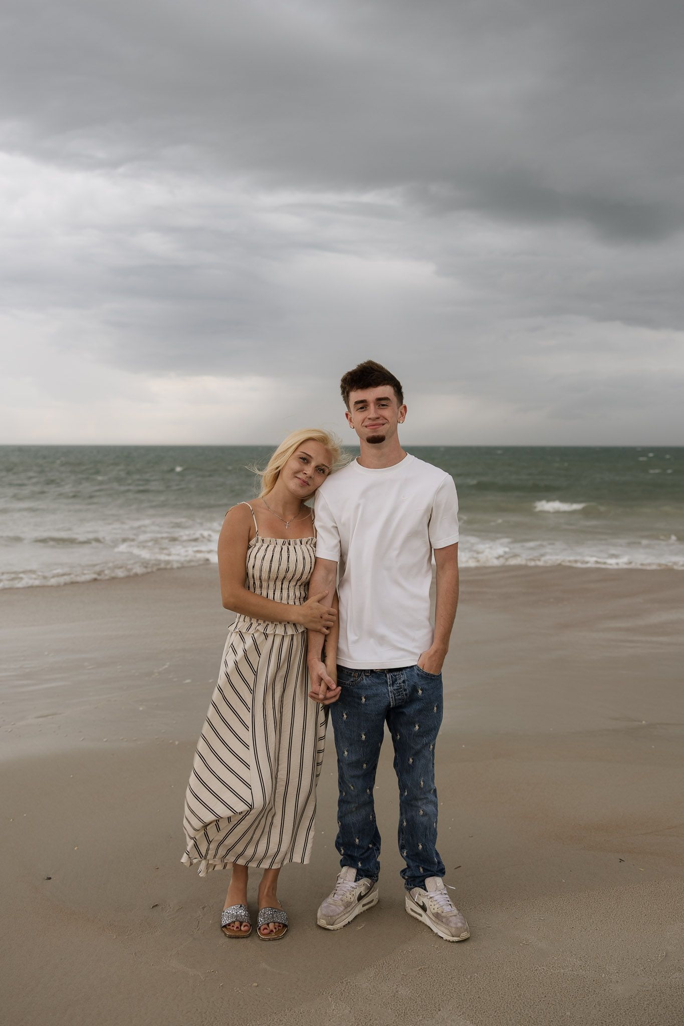 A young couple poses on a sandy beach with ocean waves and cloudy skies behind them.