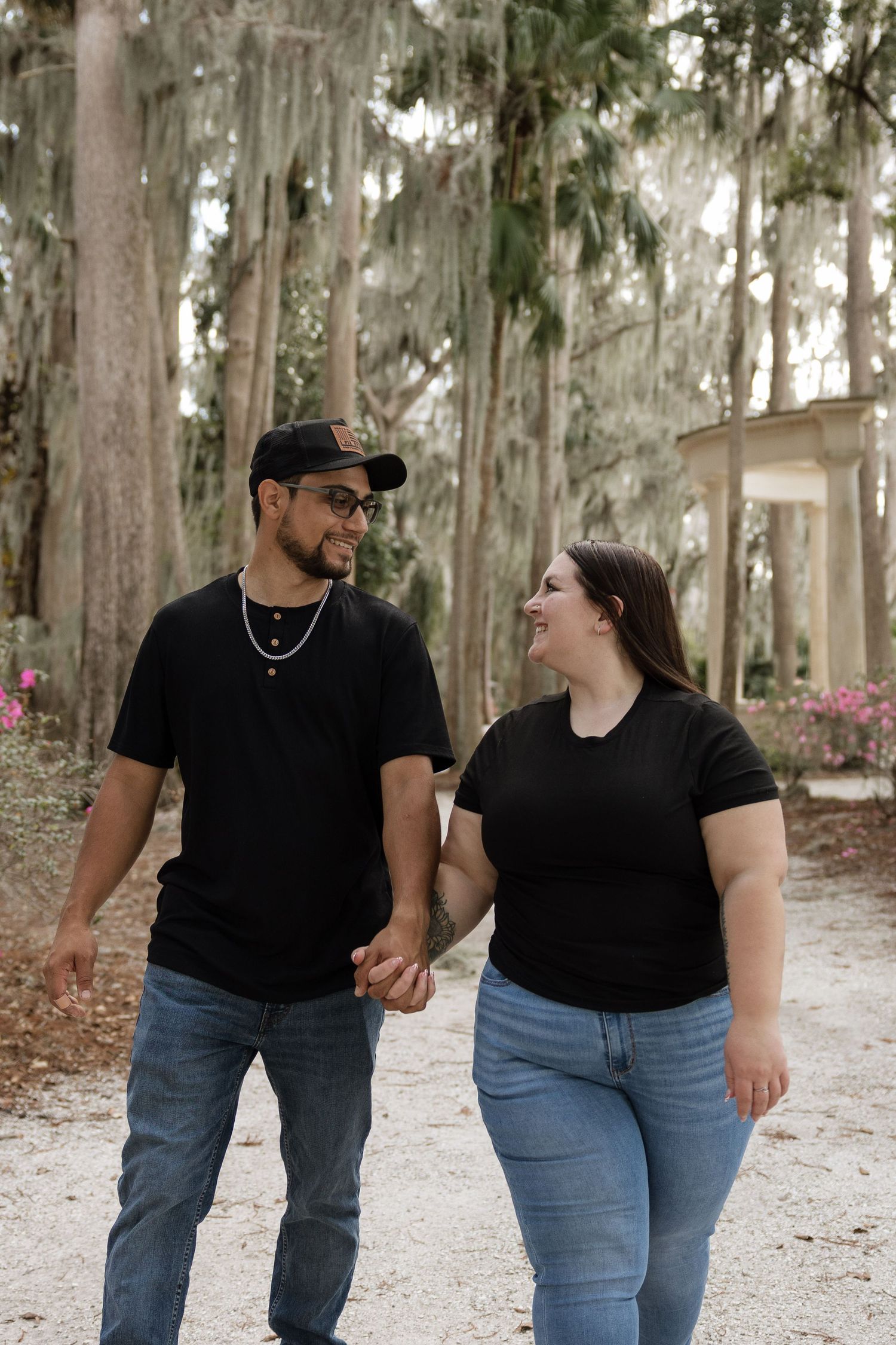 A couple wearing matching black tops and blue jeans holds hands while walking through a wooded path with Spanish moss.