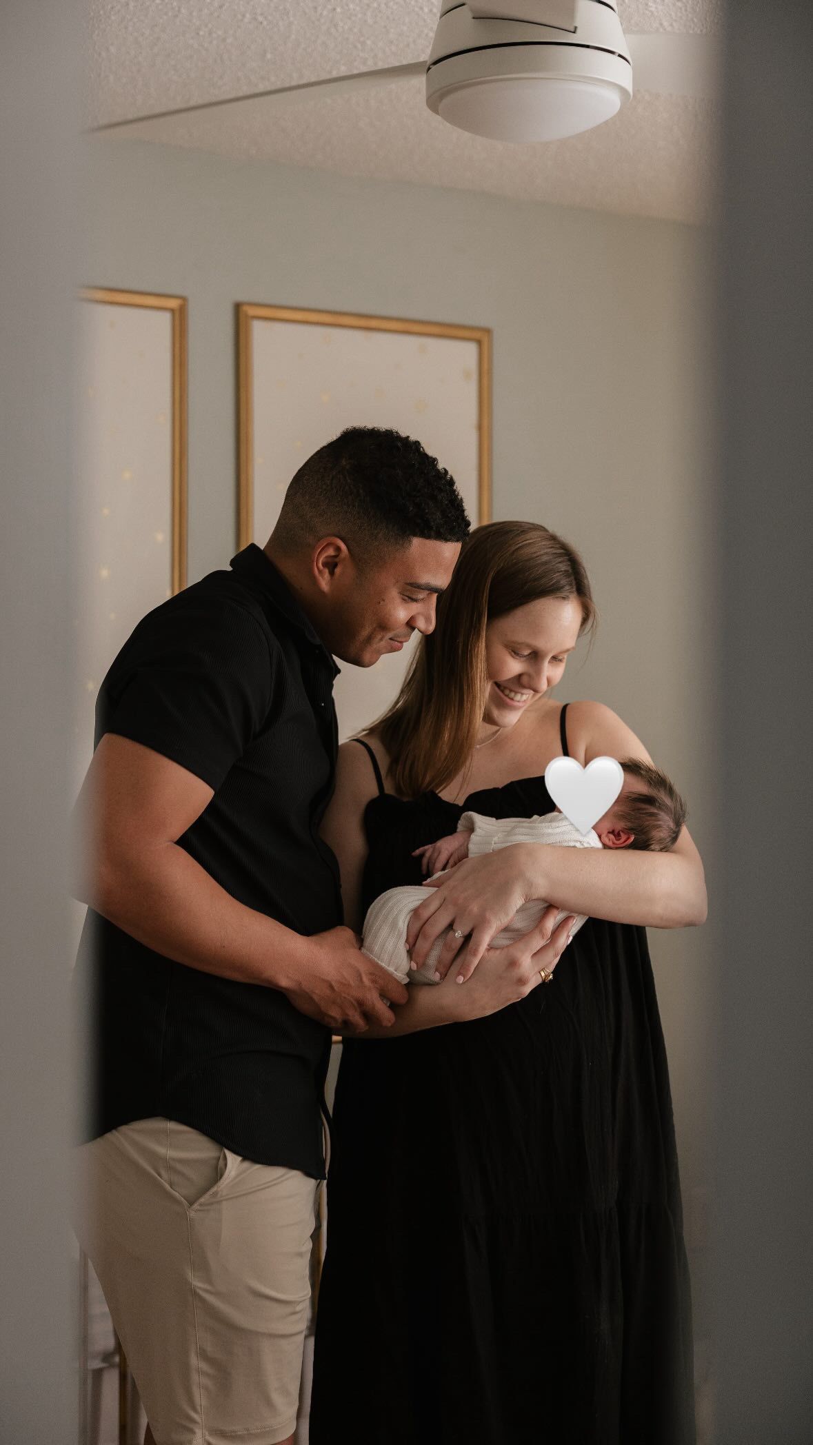 A couple in black and beige attire tenderly hold their newborn baby in a softly lit room.