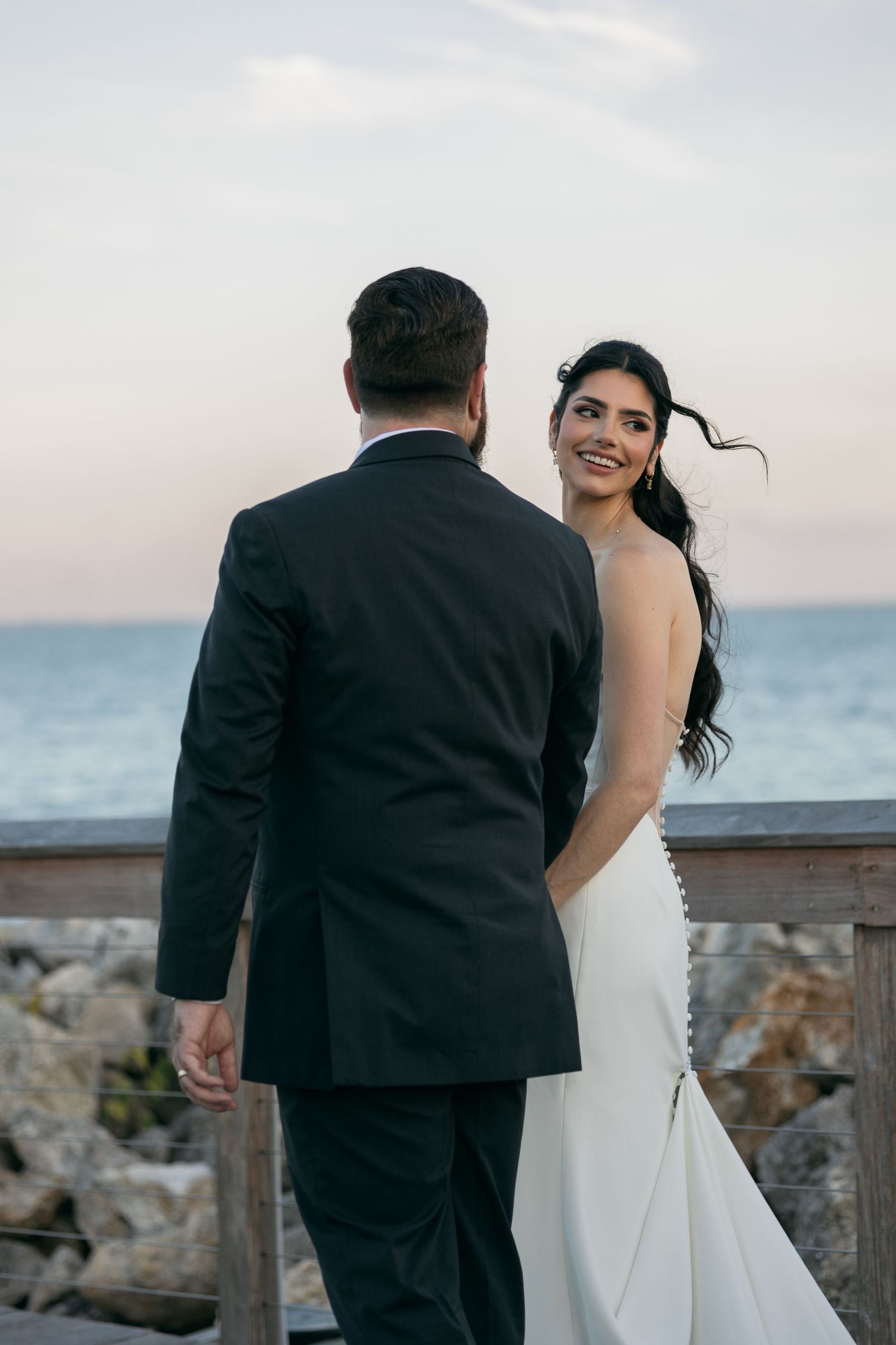 Bride in white gown smiles over shoulder at groom in black suit on seaside deck at sunset.