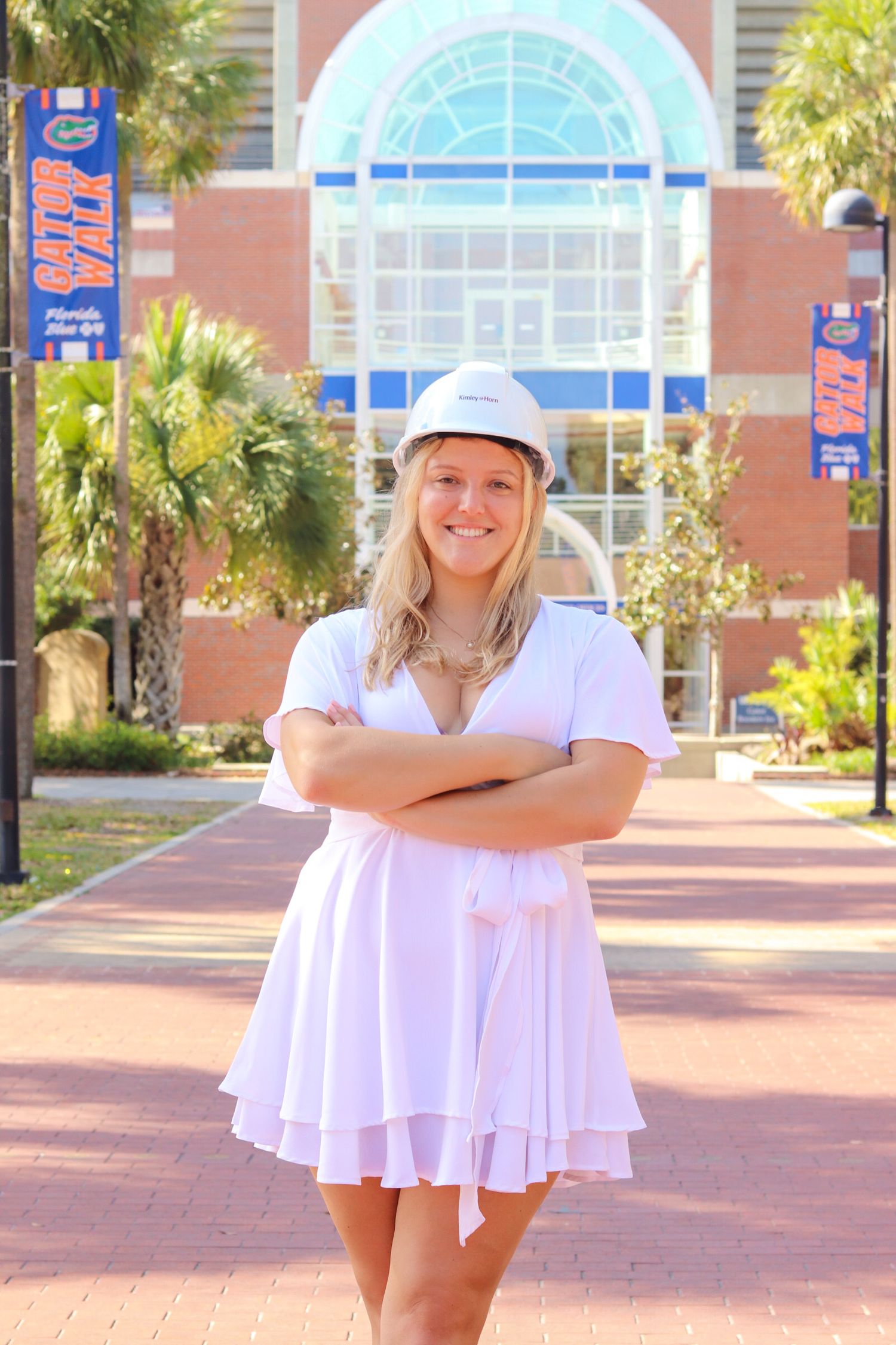 Student in white dress poses in front of university building with palm trees.