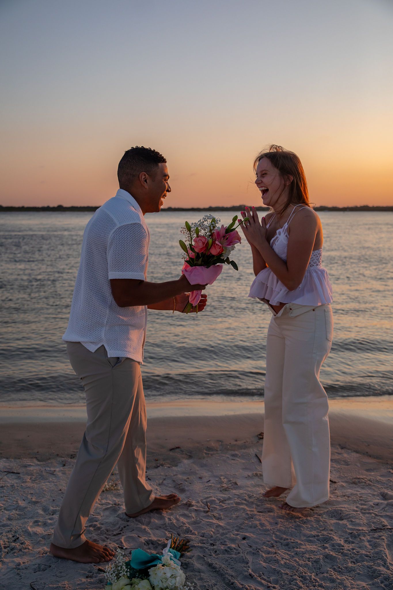 Couple exchanges romantic gestures at sunset on beach with ocean backdrop.