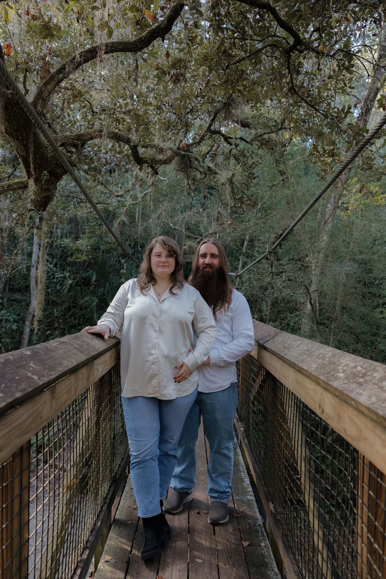 Couple in casual white outfits standing together on wooden boardwalk surrounded by trees.