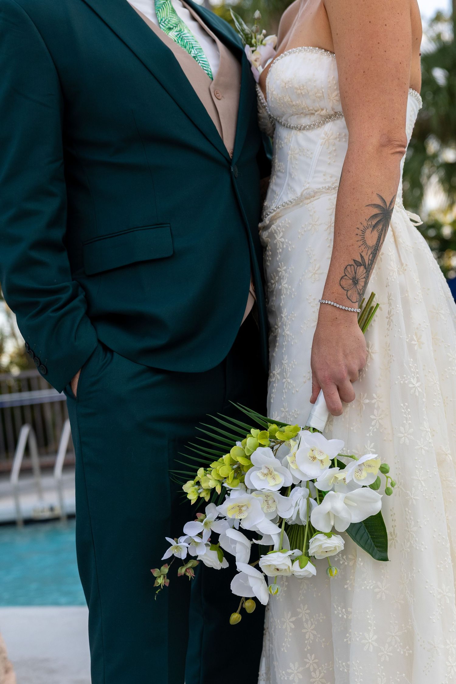 Close-up of wedding attire showing dark suit and white lace dress with orchid bouquet.