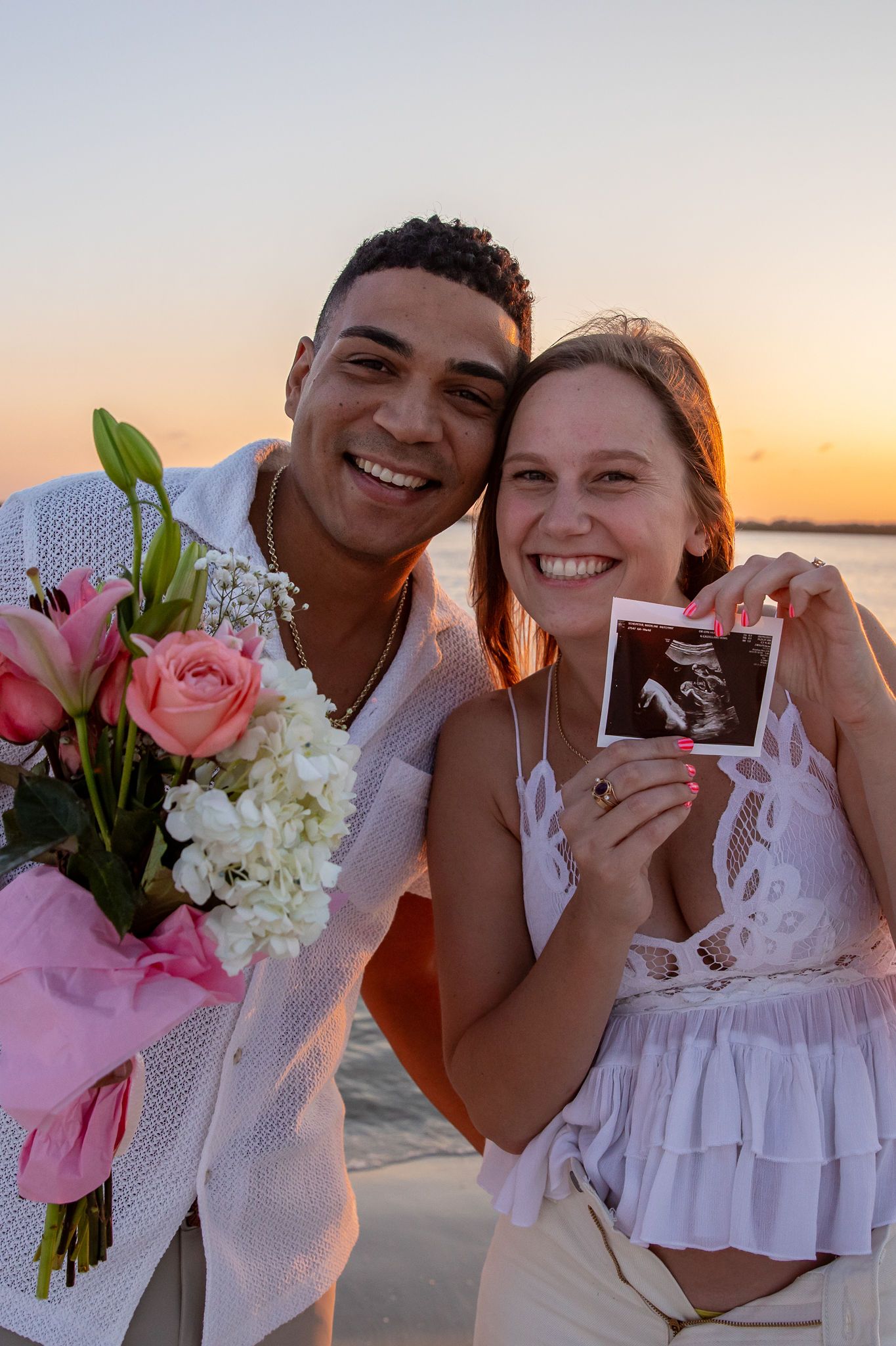 Couple celebrates on beach with flowers and ultrasound photo at sunset.
