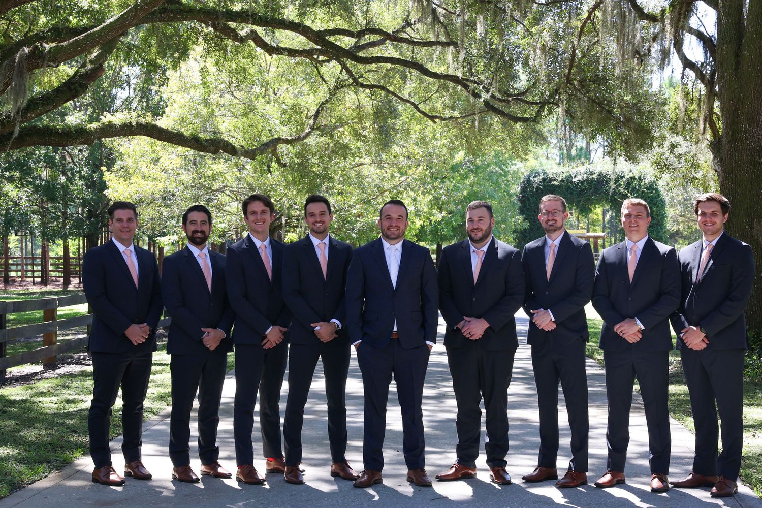 Wedding party groomsmen lined up under oak trees wearing dark suits.