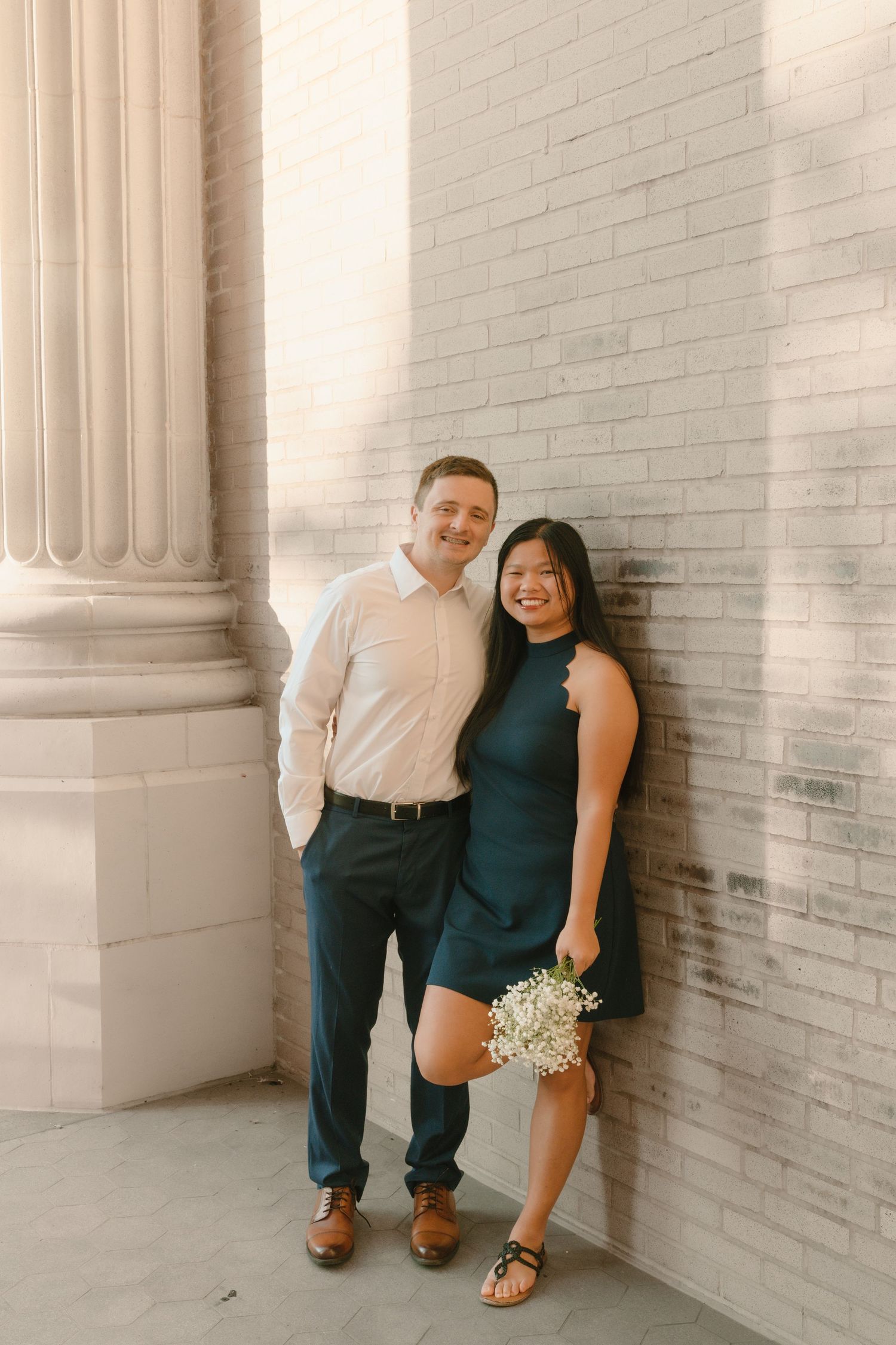 A couple poses against white brick walls and columns in formal attire.