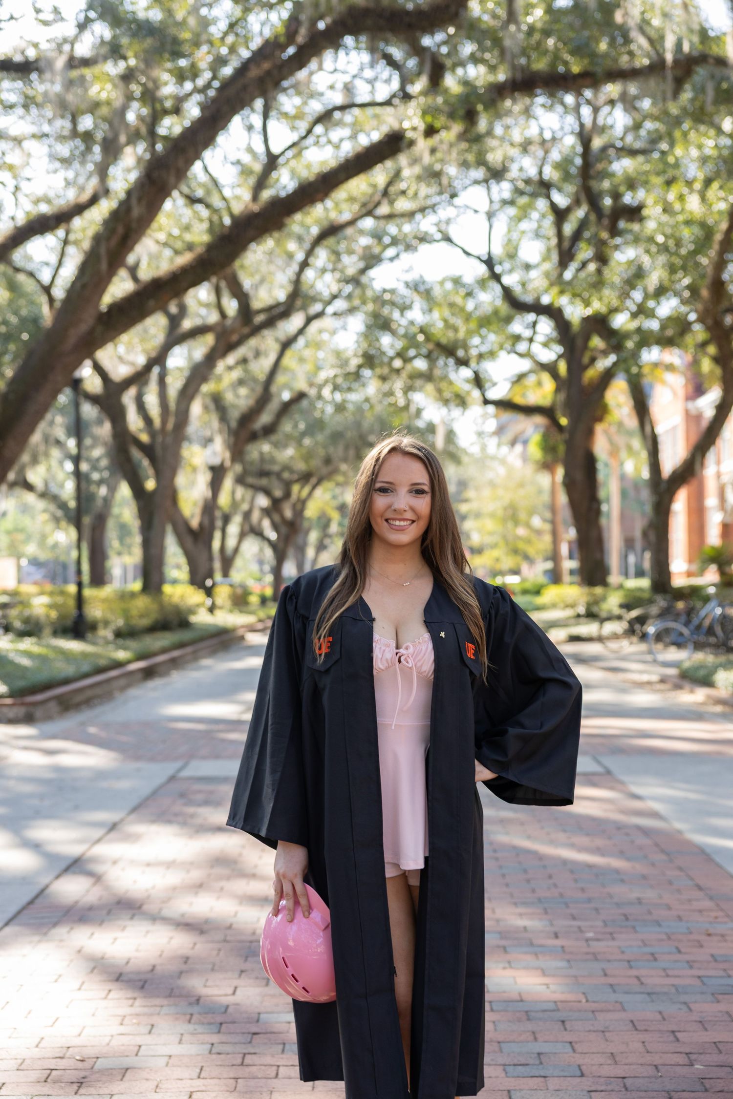 Graduate in black cap and gown poses on brick pathway lined with oak trees.