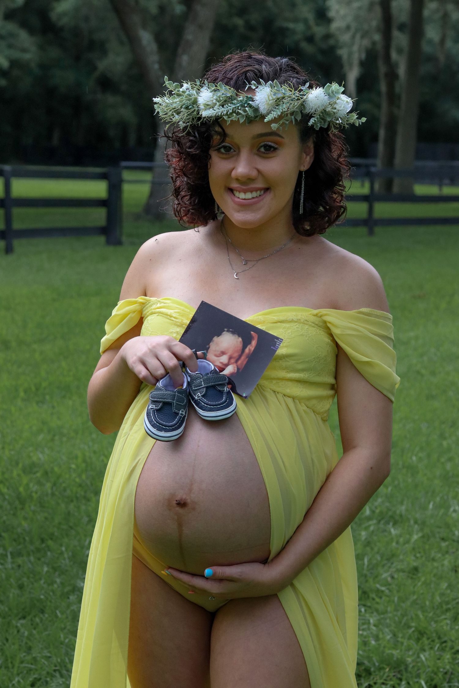 Expectant mother in yellow dress and floral crown holds tiny baby shoes in outdoor maternity photo.