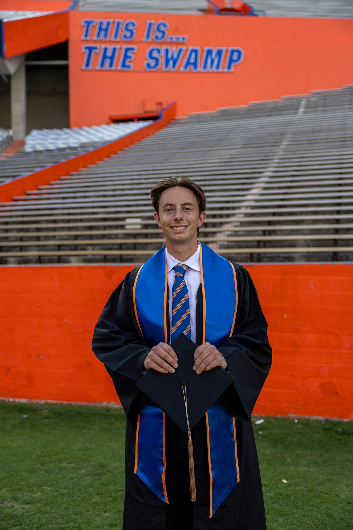 University of Florida graduate in cap and gown smiles at The Swamp stadium, wearing blue and orange stole.