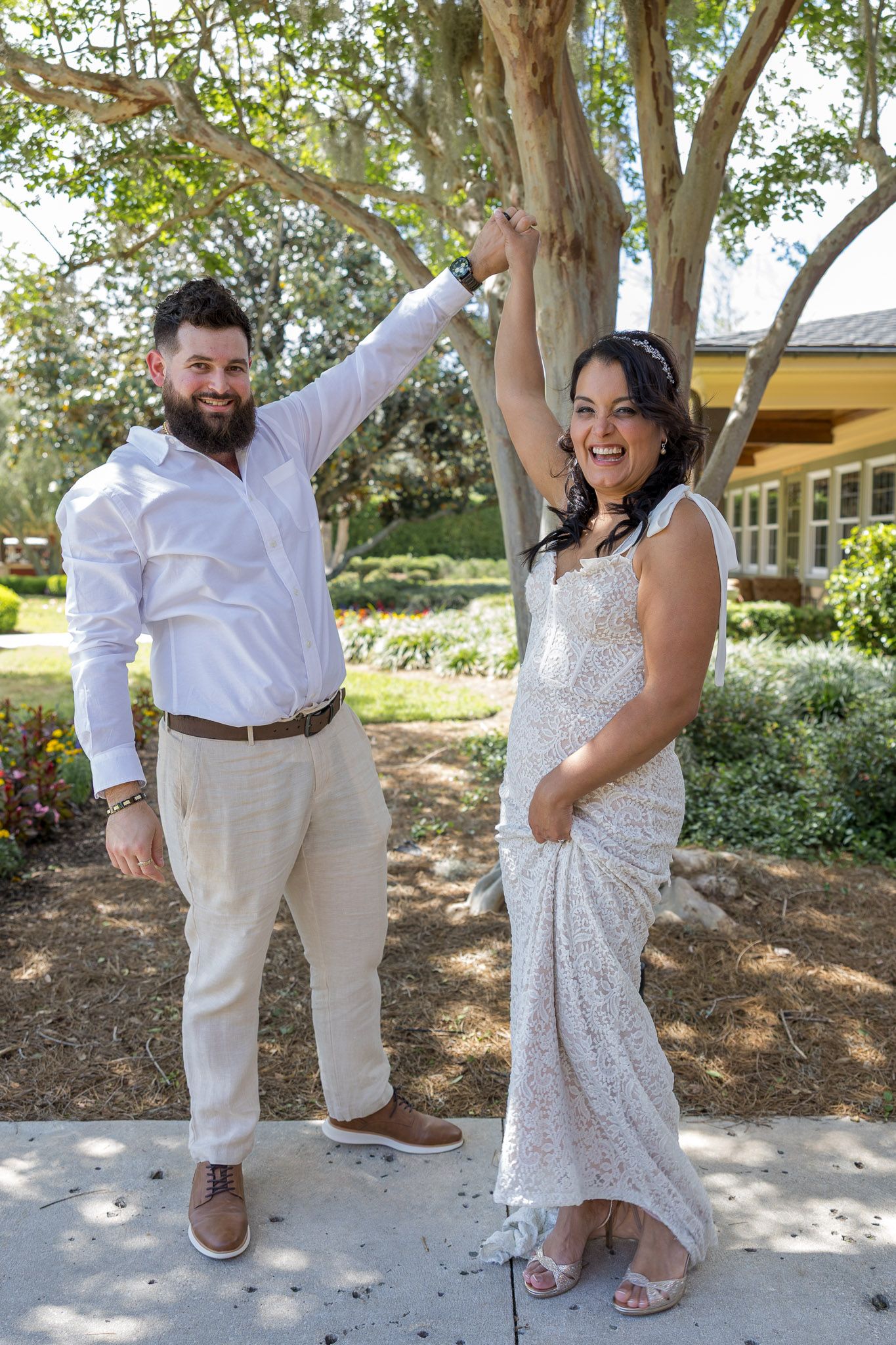Joyful newlyweds pose outdoors, groom in white shirt twirling bride in lace wedding gown under a tree.