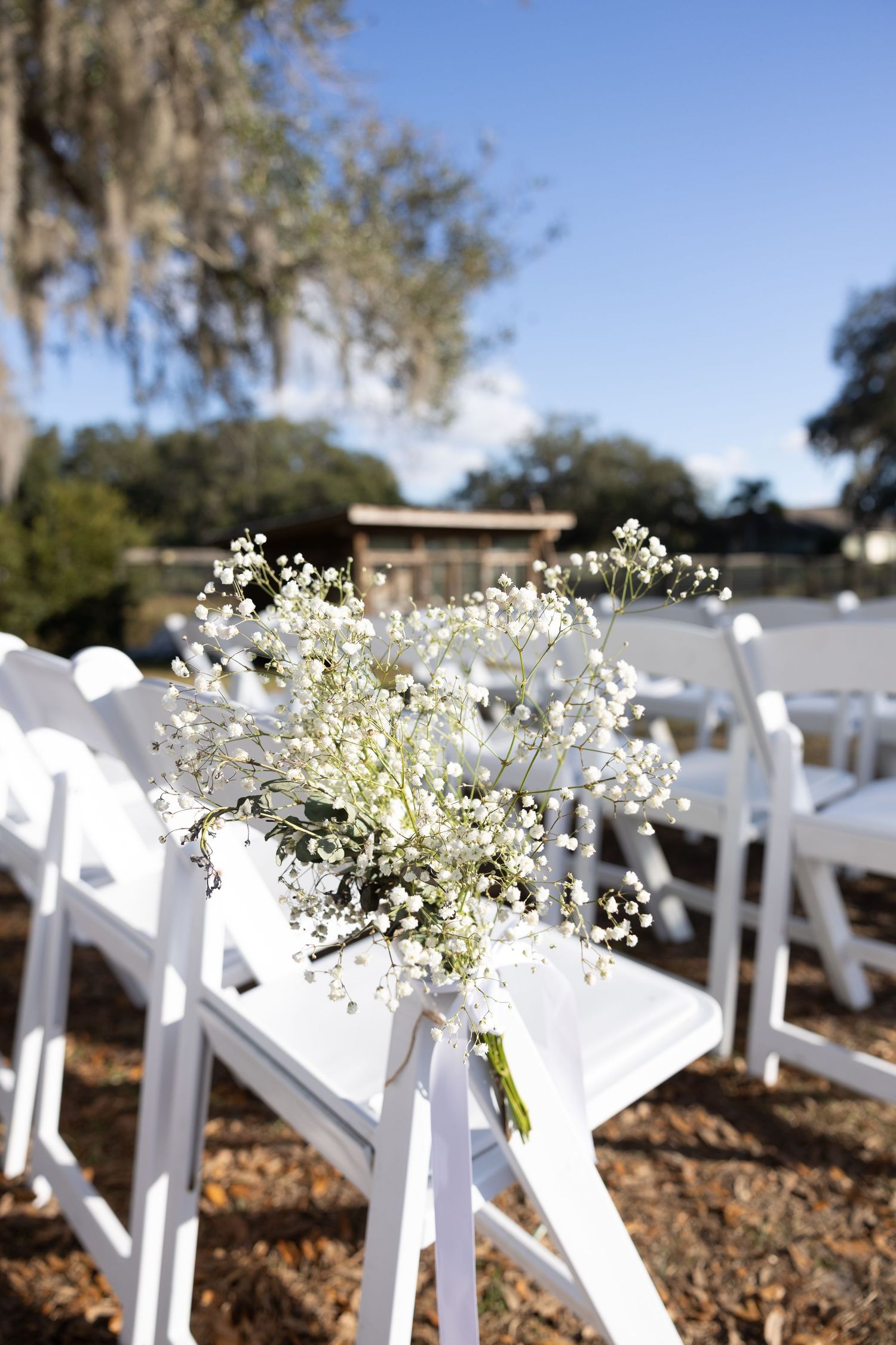 White chairs decorated with baby's breath flowers line an outdoor wedding ceremony aisle.