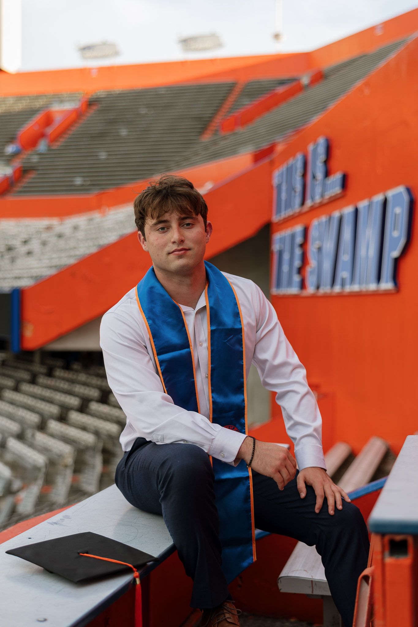 Graduate in blue sash poses at The Swamp stadium, University of Florida, with cap beside him.