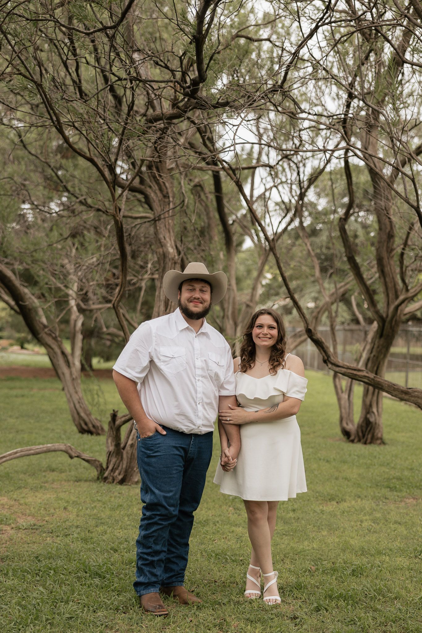 Couple posing outdoors in rustic setting, man in cowboy hat and jeans, woman in white dress, surrounded by trees.