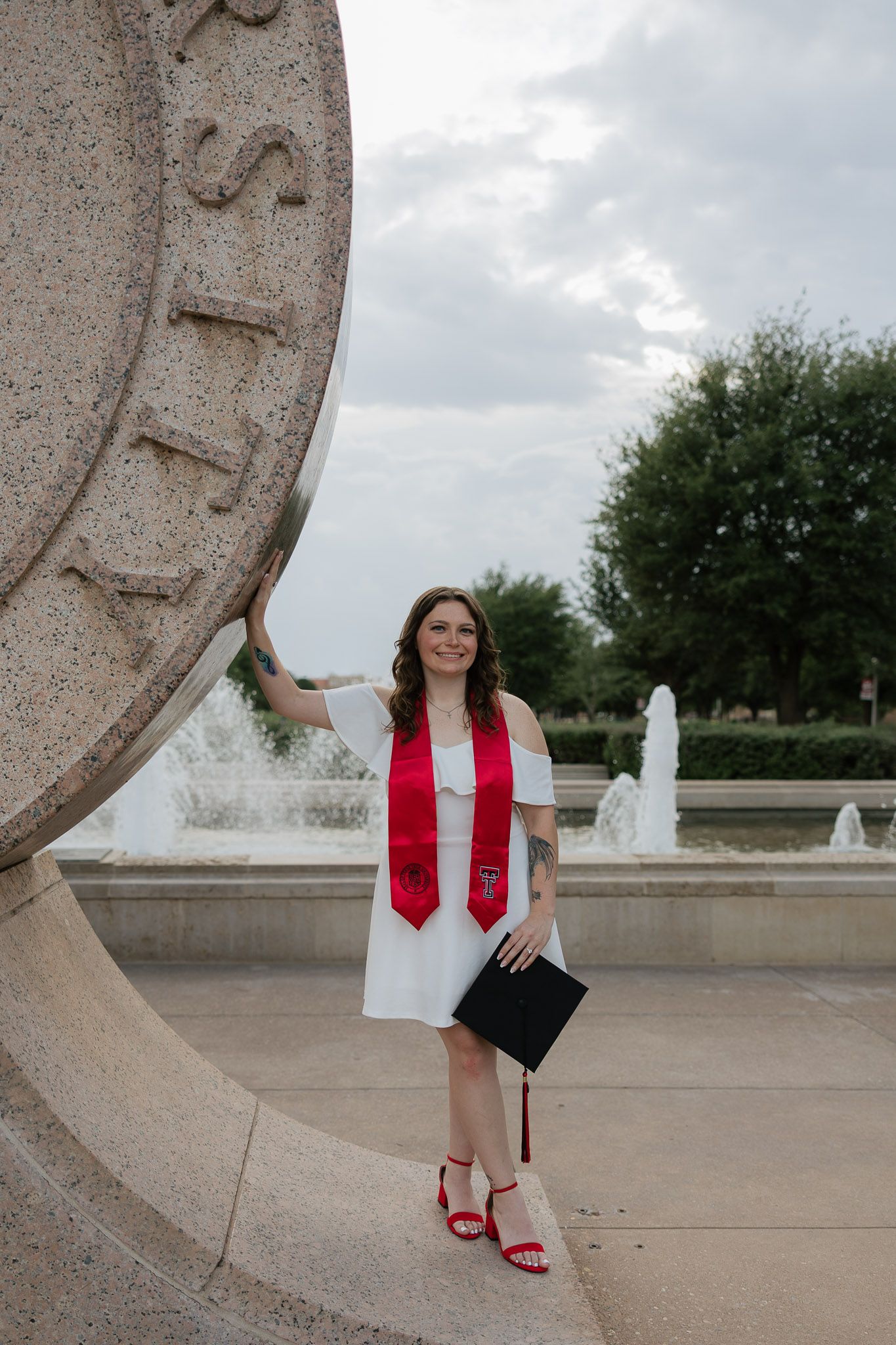 Smiling graduate in white dress and red stole holds cap beside university seal with fountain backdrop.