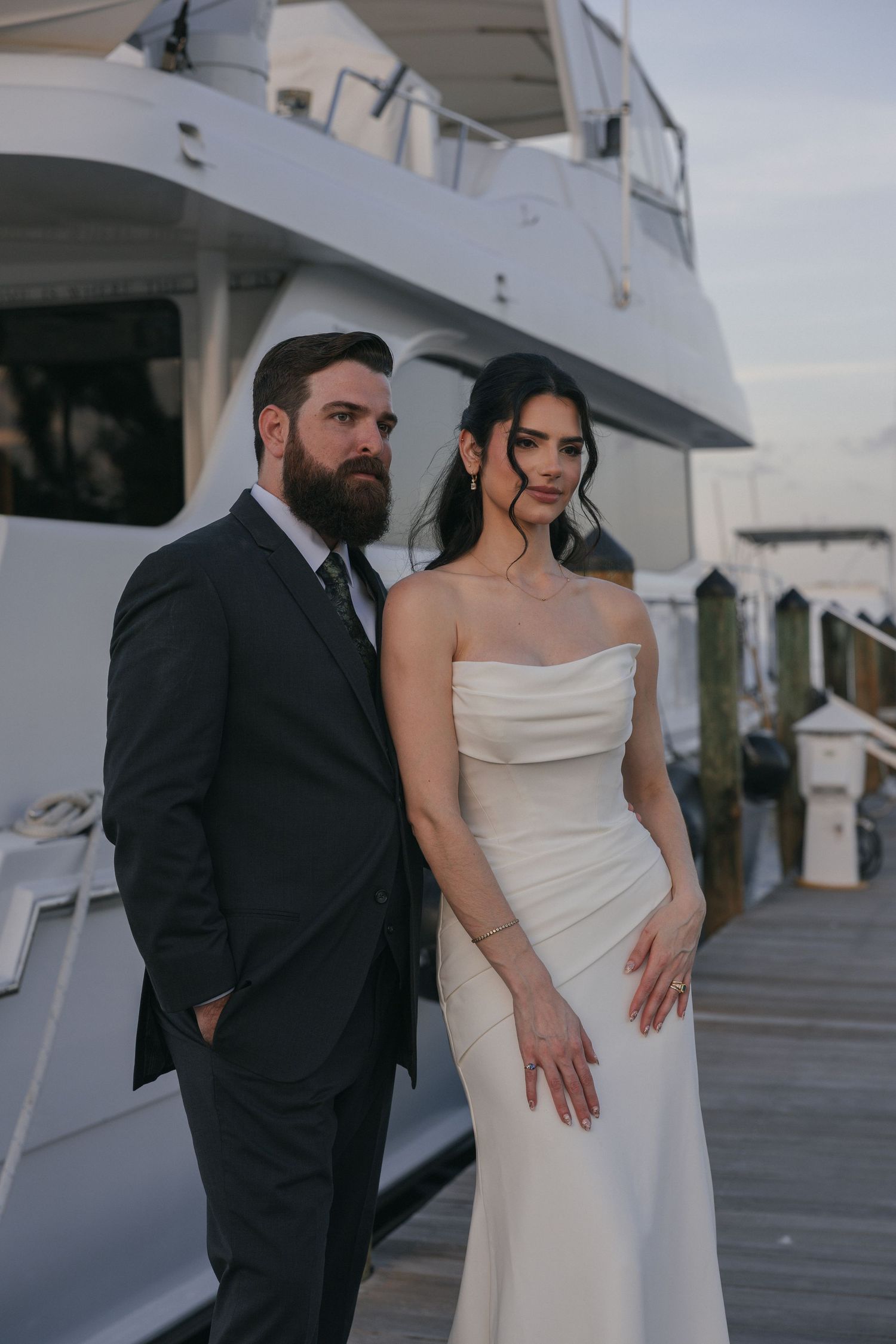 Bride in strapless white gown and bearded groom in dark suit pose on marina dock near yacht.