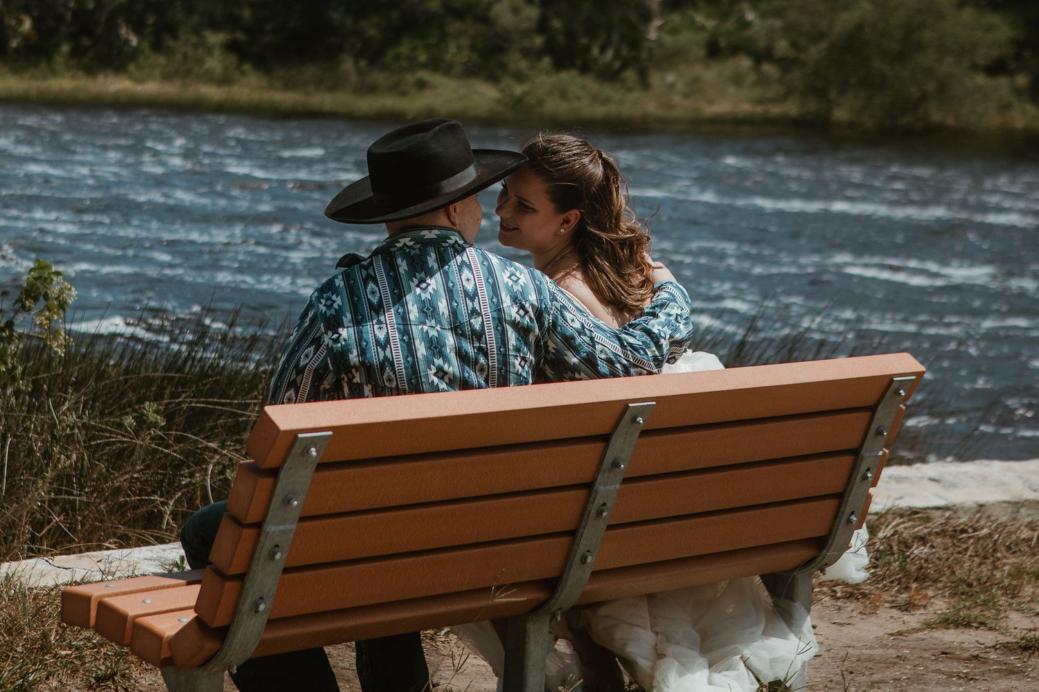A couple sits together on a wooden bench overlooking a scenic river.