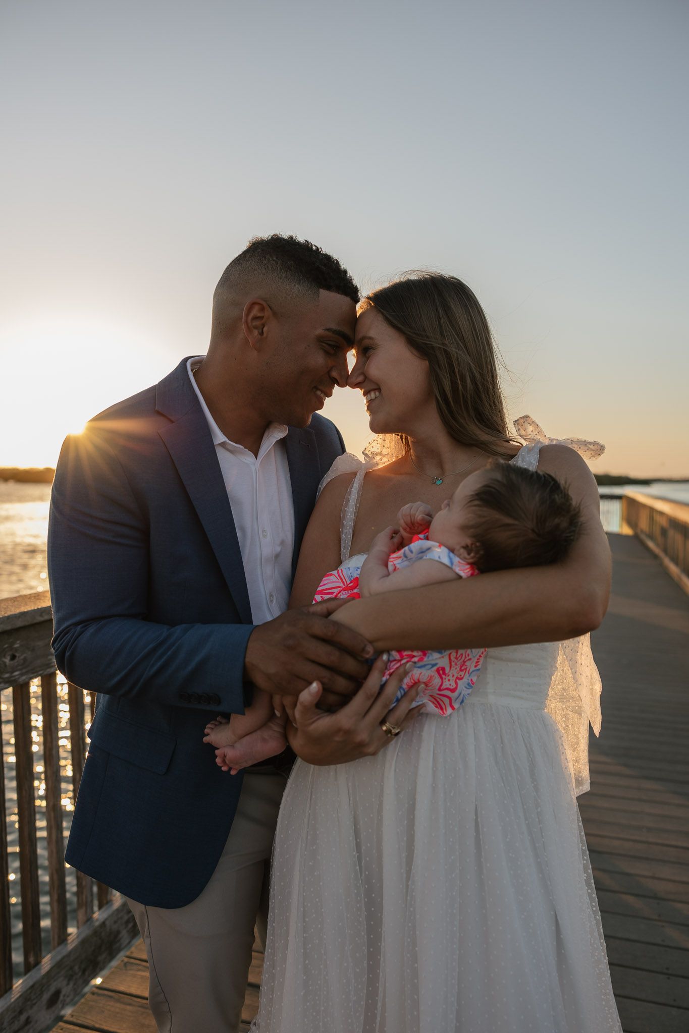 Couple sharing a romantic sunset moment on a pier while tenderly holding their newborn baby dressed in pink.