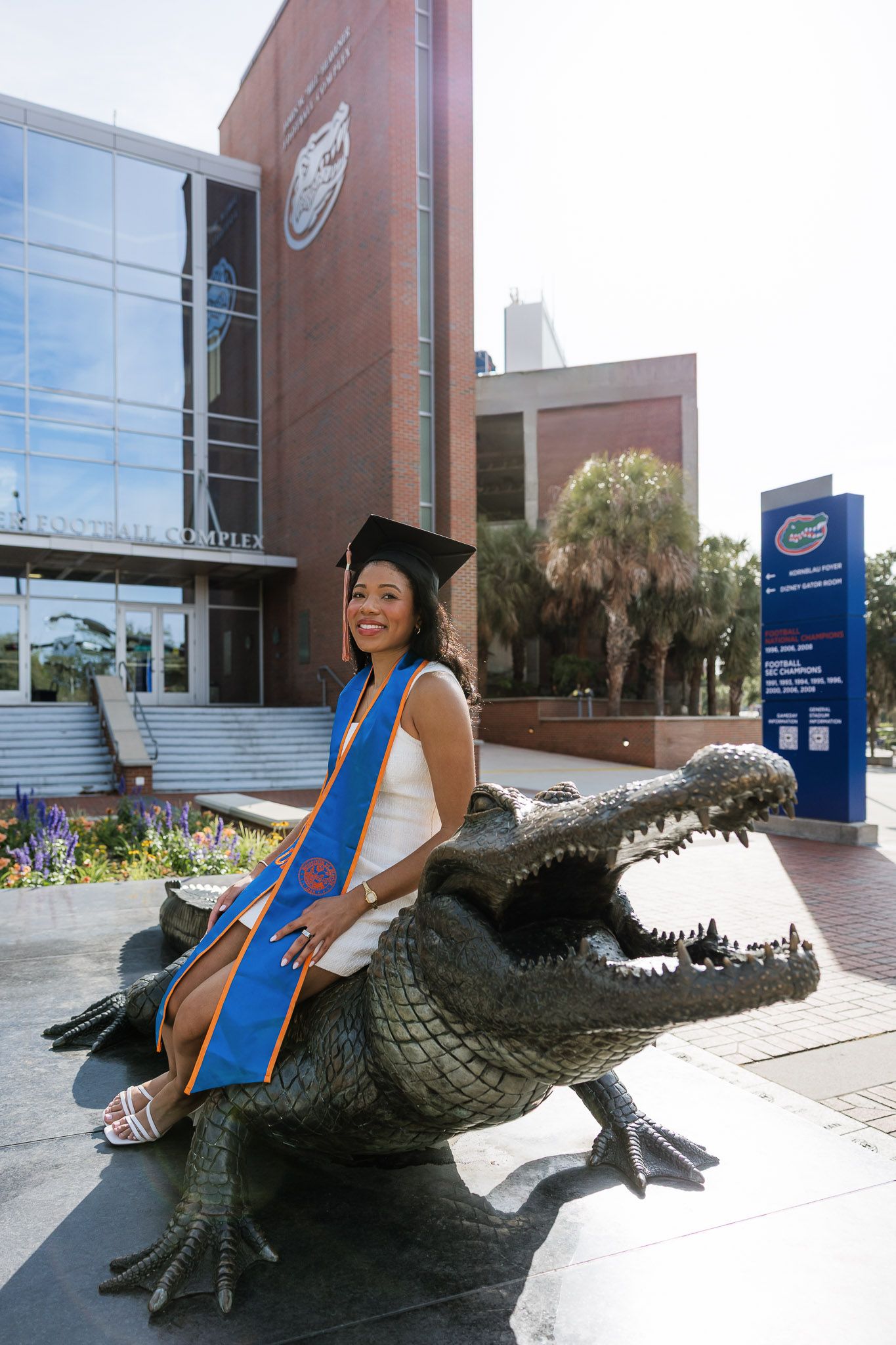 Graduate in cap and gown sits on alligator statue at University of Florida, smiling in front of campus building.