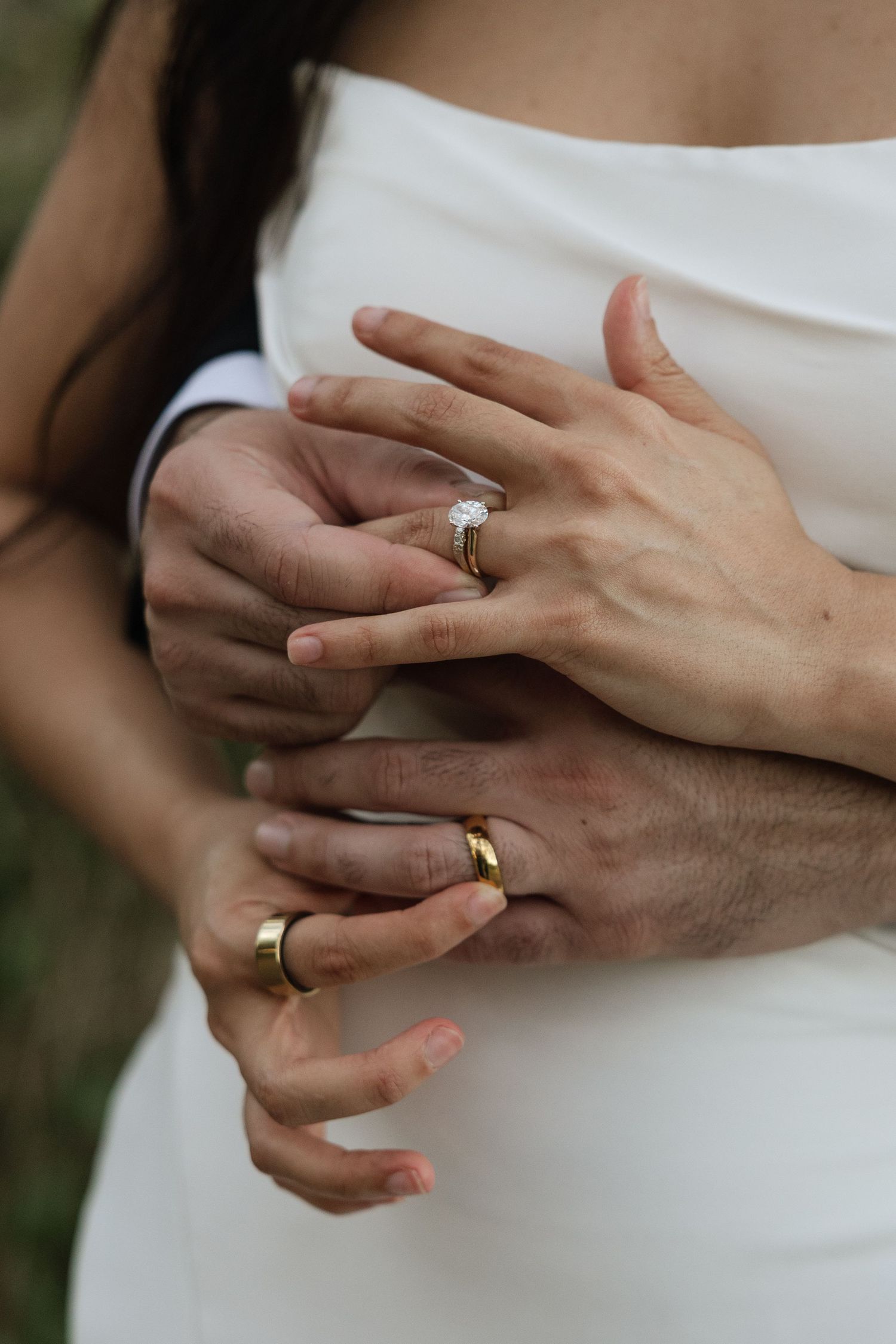 Close-up of couple's hands with wedding rings embracing, showing engagement ring and gold bands against white dress.