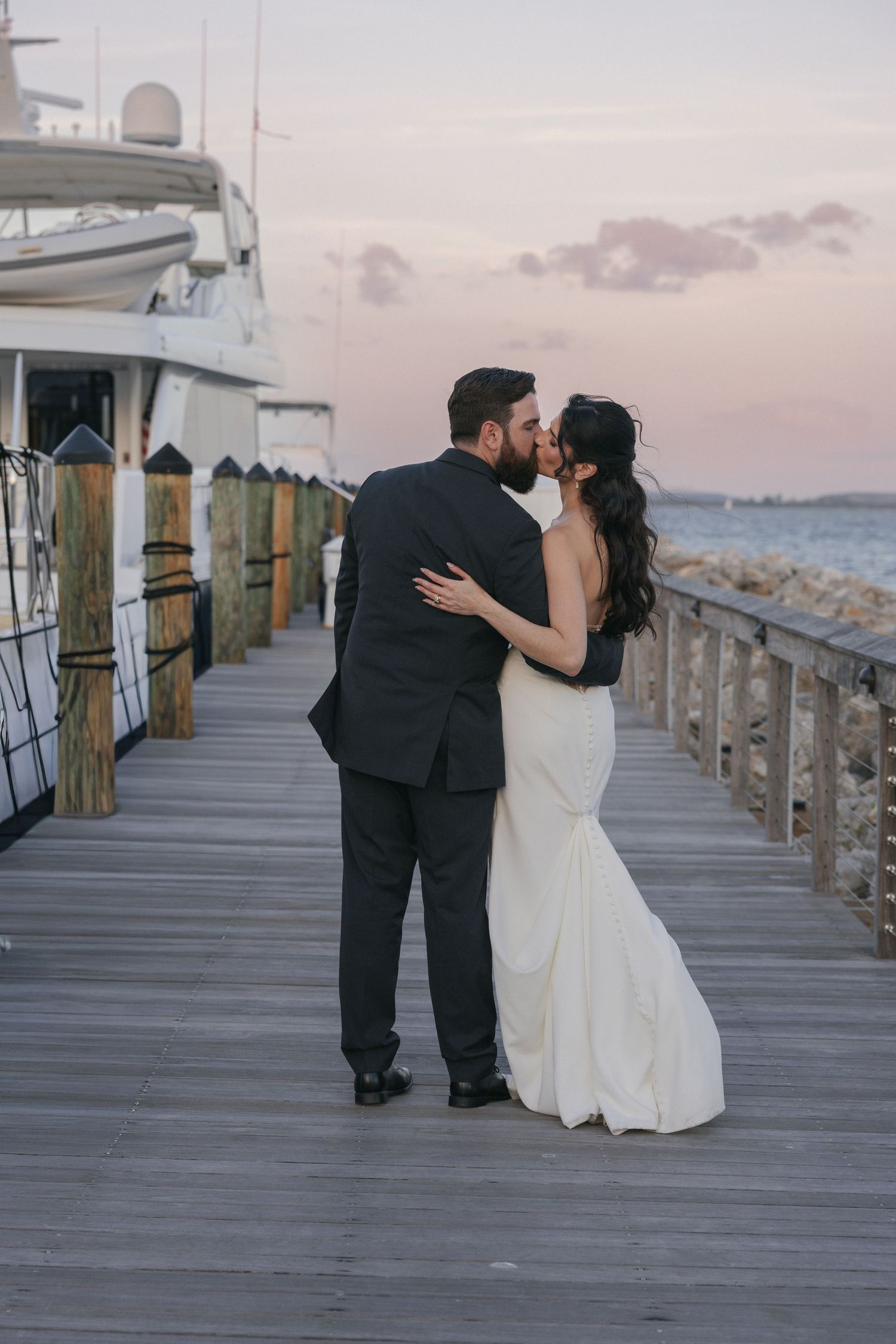 Bride and groom share a romantic kiss on a waterfront dock at sunset, with a yacht and pastel sky in the background.