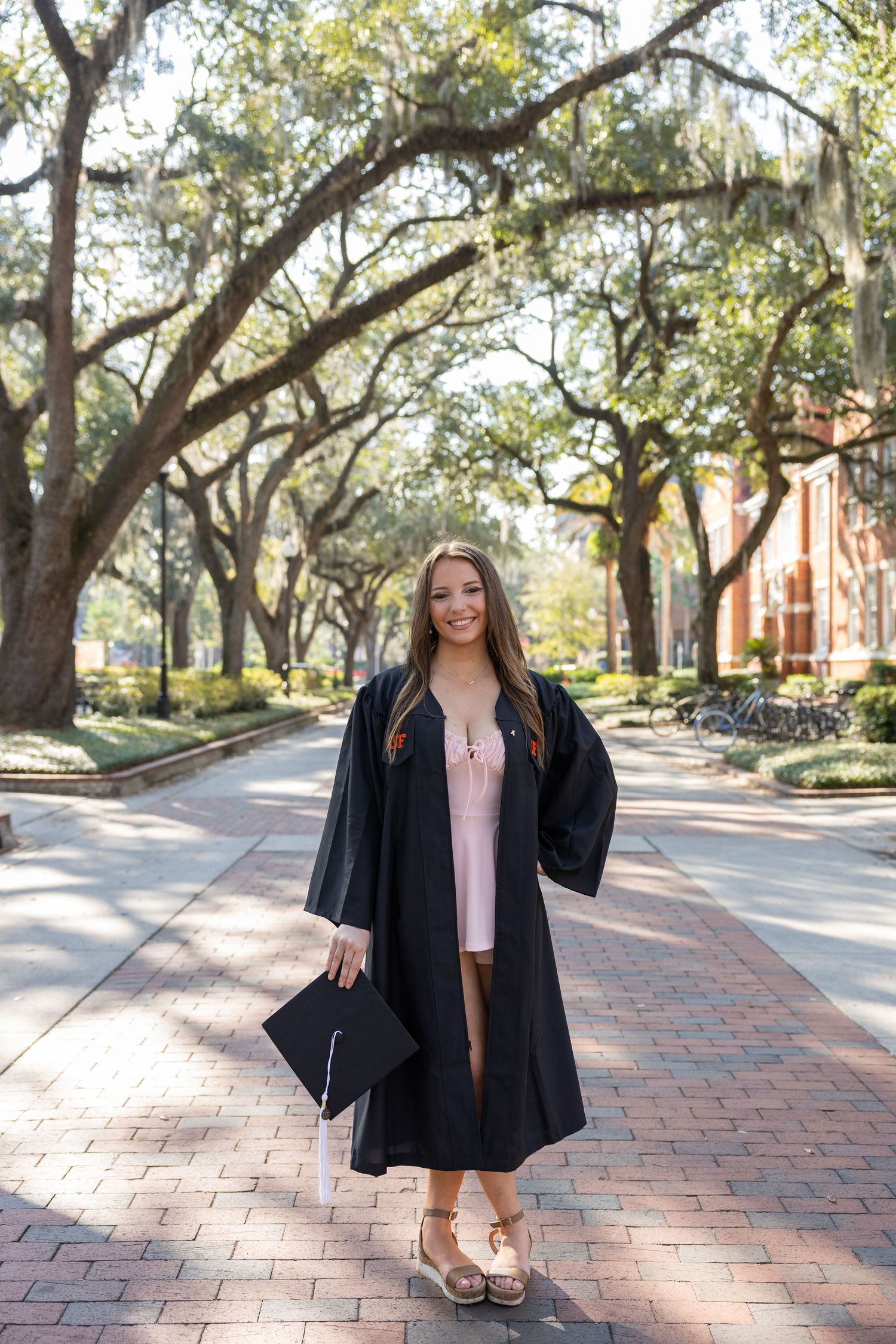 Graduate in black gown poses on brick pathway under canopy of oak trees.