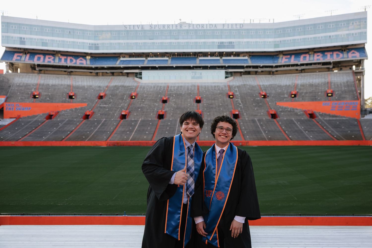 Two graduates in caps and gowns pose proudly at Florida's Ben Hill Griffin Stadium on graduation day.