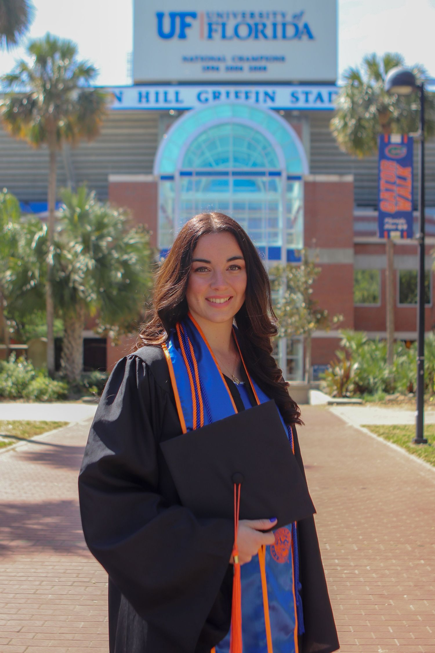 Graduate in cap and gown poses in front of Ben Hill Griffin Stadium at University of Florida.